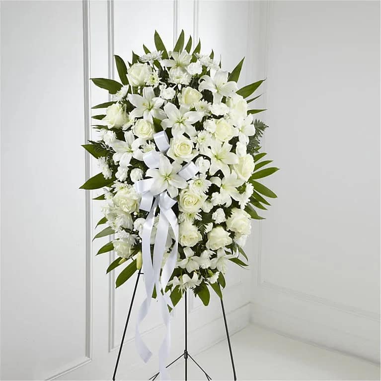 Blooming Joy - “Pay tribute to a cherished life with this elegant standing spray of pure white flowers. Featuring majestic lilies, delicate roses, carnations, and chrysanthemums, this arrangement is accented with lush greenery and adorned with a graceful white ribbon. Perfect for conveying peace, love, and remembrance, this timeless design honors and celebrates a life well-lived. ”