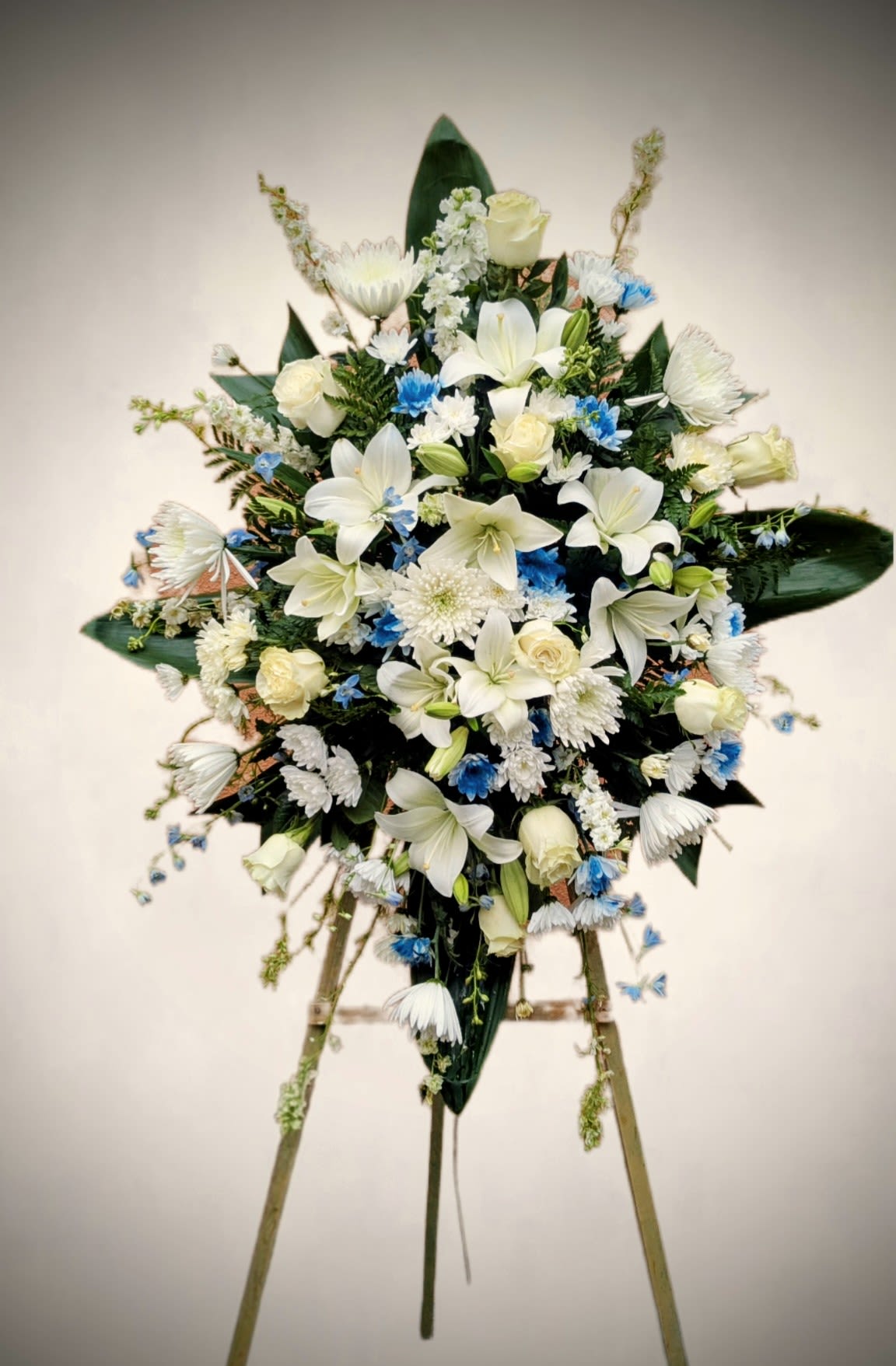 Elegant White and Blue Standing Sympathy spray  - Honor a loved one with this beautifully crafted standing sympathy spray, designed to convey peace, comfort, and heartfelt remembrance. This arrangement features a lush blend of white lilies, roses, chrysanthemums, and carnations, accented with delicate blue blooms for a serene touch of color. Set against rich greenery and presented on a sturdy easel, it creates an elegant display suitable for funeral services, memorials, or graveside tributes.  Thoughtfully arranged to symbolize purity, tranquility, and eternal love, this standing spray offers a graceful way to express your condolences and support.  