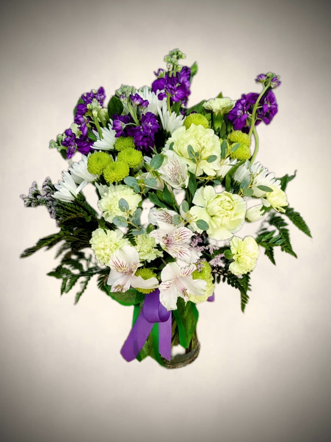 Lavender and Lime Fresh Blooms  - Bright, cheerful, and full of charm, this vase arrangement combines soft elegance with vibrant color. Featuring a mix of creamy white carnations, delicate alstroemeria, lime-green button mums, and deep purple stock flowers, this bouquet offers a refreshing blend of textures and tones. Fresh greenery adds natural fullness, while a coordinating purple ribbon accent completes the look with a graceful touch.  Perfect for birthdays, get-well wishes, thank-you gifts, or simply brightening someone’s day, this arrangement brings uplifting beauty to any space. 