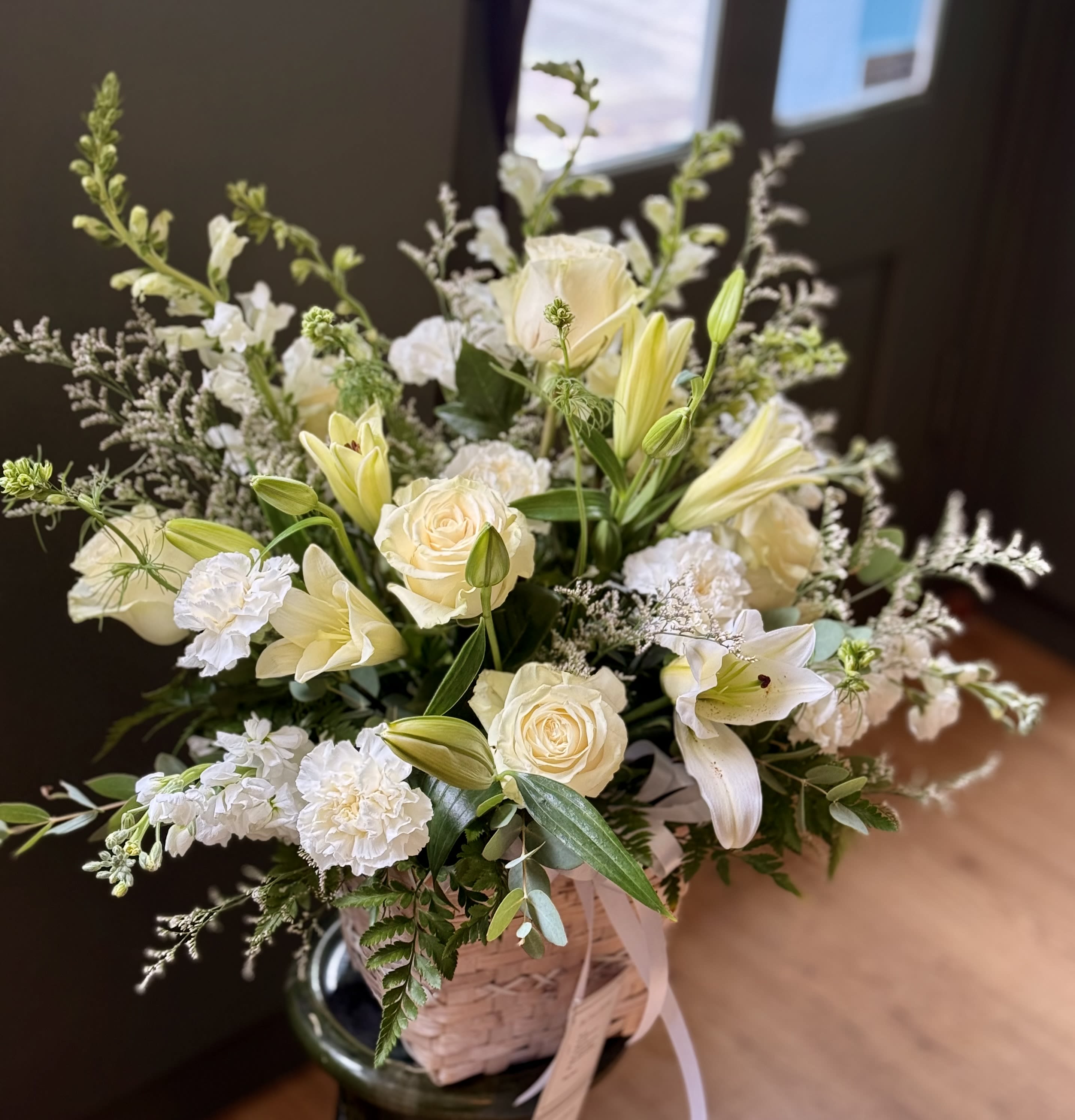 Serenity Basket - Beautiful basket with white flowers including roses, lilies, carnations, Astros, Stock, Snaps, with greenery. Basket size: 6&quot; H 9 1/2&quot; opening. Arrangement approximate size including basket:  17&quot; H 18&quot; D