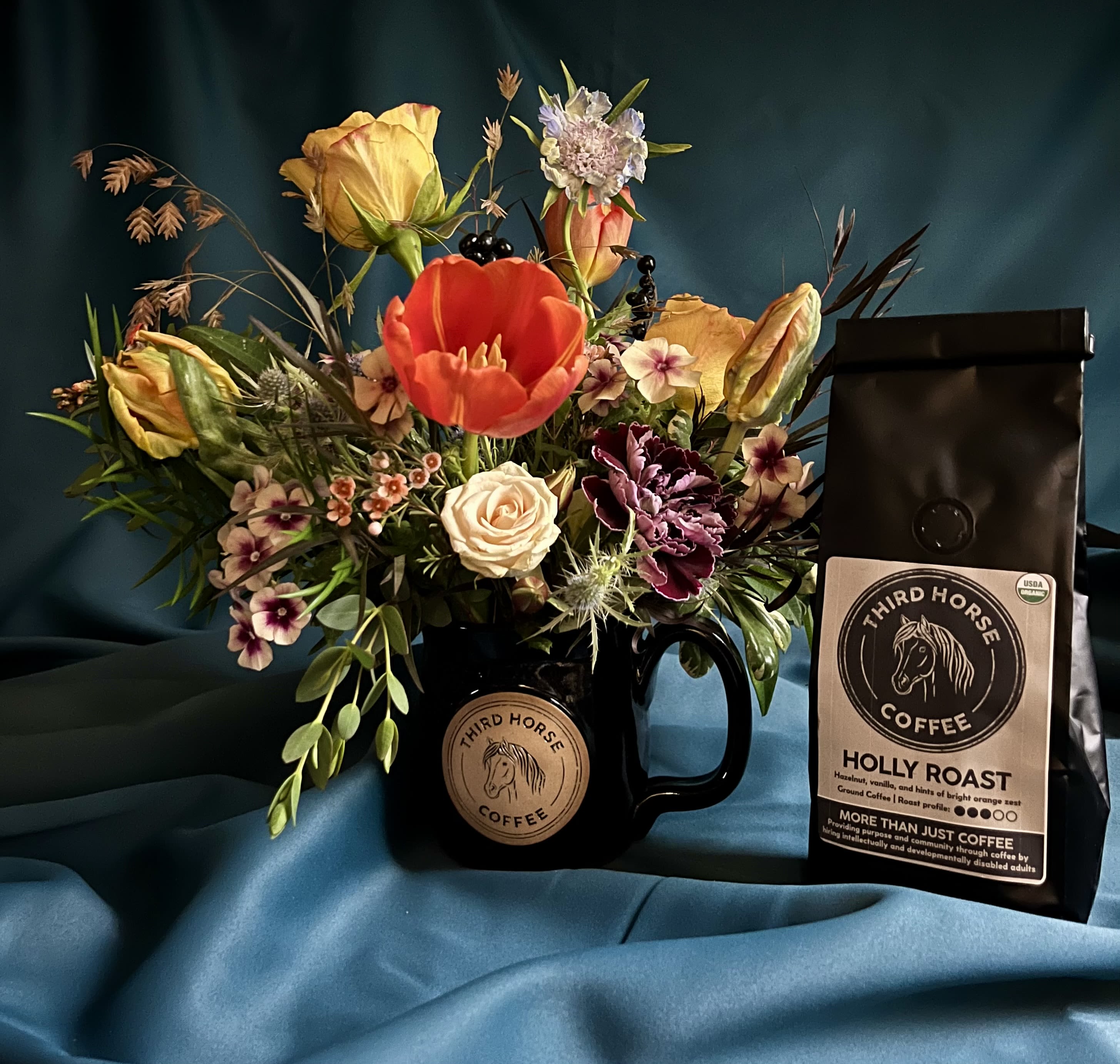 FLOWER AND COFFEE GIFT PACK - For the coffee lovers! The Third Horse Mug and Coffee arrangement is a delightful choice that showcases our appreciation for Third Horse Coffee. Priced at $75, this handsome arrangement includes a hefty, reusable mug and a bag of Third Horse Organic Ground Coffee, featuring their special holiday roast, Holly Roast. This blend offers delicious flavors of hazelnut, vanilla, and a hint of bright orange zest.  In addition to the Holly Roast, you can choose from other enticing flavors: the Wild Stallion (Dark) roast delivers a full-bodied experience with notes of citrus, caramel, and a smoky finish, while the Guatemalan (Medium) roast features delightful hints of brown sugar, maple, and fig with a smooth finish. Please specify your preferred roast type when placing your order. By purchasing this arrangement, you’re also supporting Third Horse Coffee, a non-profit specialty coffee shop dedicated to hiring adults with intellectual and developmental disabilities, helping them gain purpose, job skills, and a sense of community.