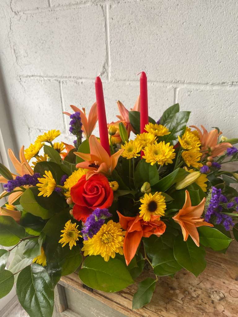 Autumn Candlelight Centerpiece - A vibrant autumn centerpiece featuring orange lilies, yellow daisies, a bold red rose, and touches of purple statice. Designed in a lush bed of greenery and finished with two classic red taper candles, this piece brings warm seasonal color to any Thanksgiving table.