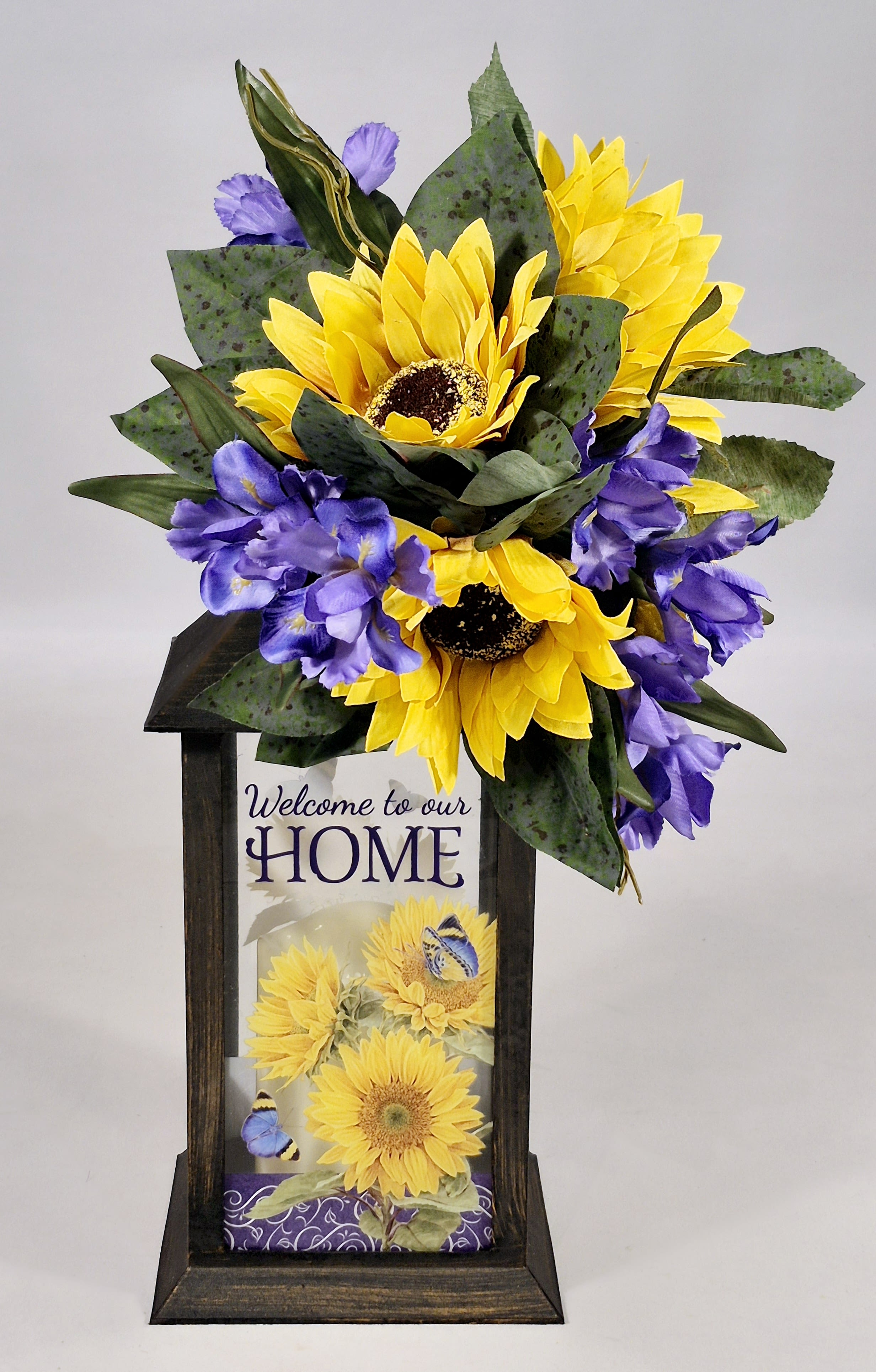 Welcome to Our Home Sweet Sunflowers Lighted Lantern - Lanterns are made of weather friendly materials and UV printed glass. They include a flame-less LED candle with a built-in automatic timer: 6hrs ON/18hrs OFF (3 AAA batteries are included). They are suitable for hanging or free-standing use. Comes with a beautiful bow and silk floral arrangement. Perfect gift for any occasion.