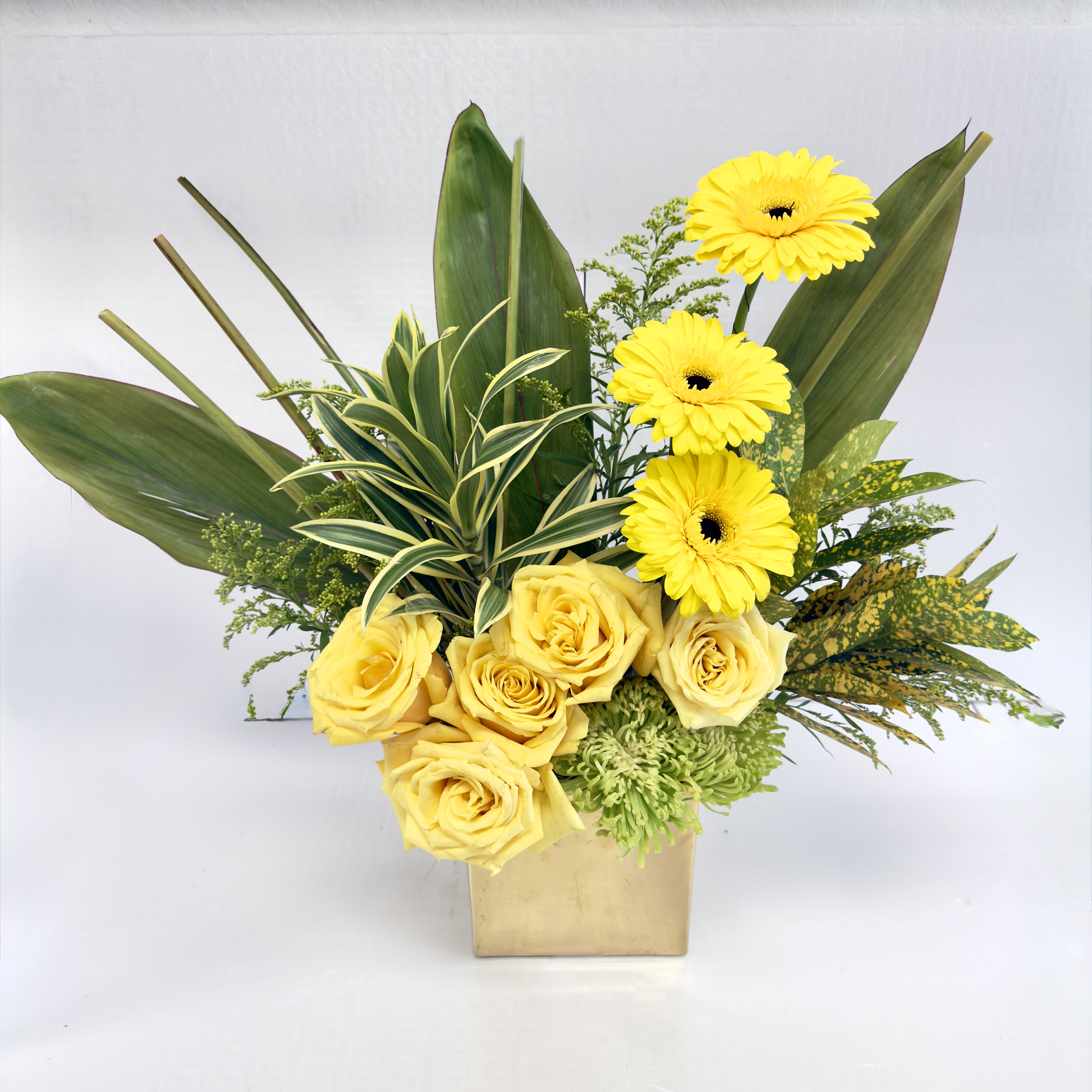 Golden Glow - Bright and uplifting, this modern arrangement features sunny yellow roses and cheerful gerbera daisies paired with lush tropical greens in a chic gold vase. A perfect choice to bring warmth, joy, and elegance to any space or occasion.