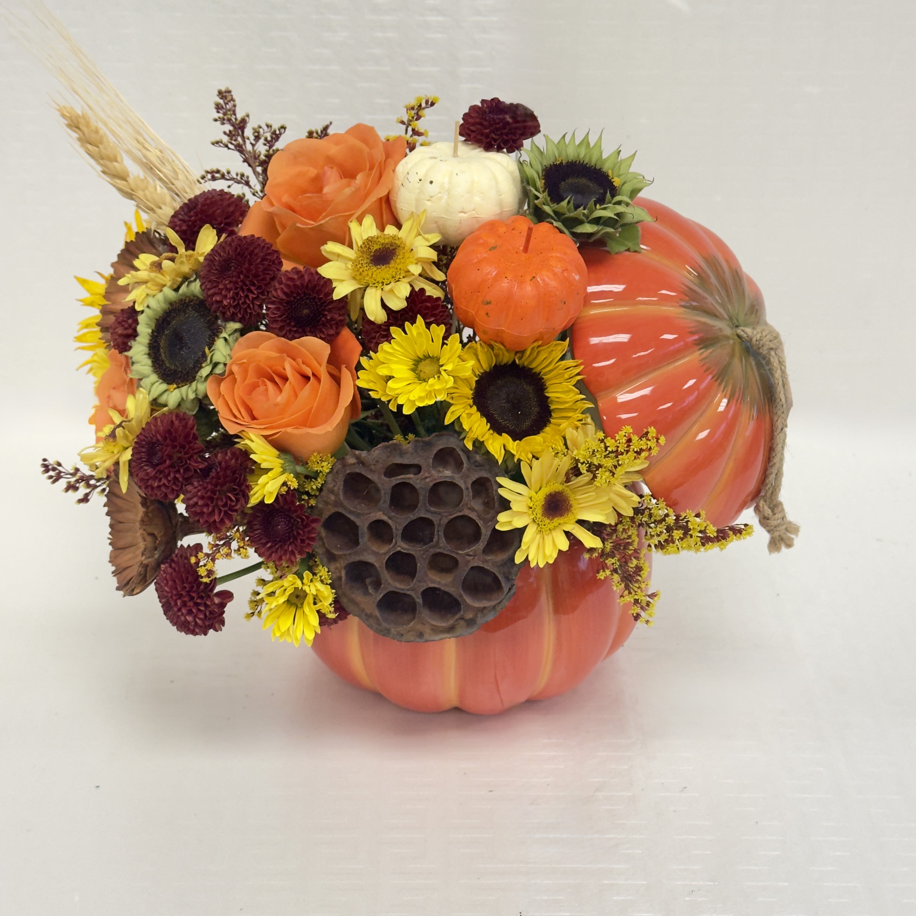 Harvest Pumpkin Bloom - A cheerful autumn arrangement featuring vibrant orange roses, sunny yellow daisies, rich burgundy mums, and rustic lotus pods, all artfully gathered in a festive pumpkin container. Perfect for brightening your home or gifting a touch of fall warmth!