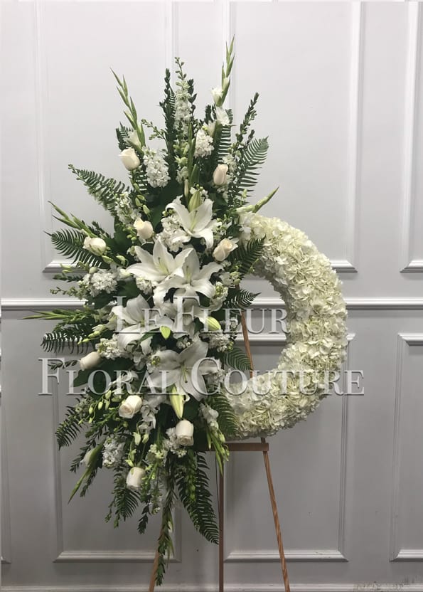White Sympathy Unique Wreath - This beautiful arrangment is; half standing spray and half sympathy wreath. With the cluster consisting of white lilies, gladiolas, stock, roses, snapdragons and delicate fillers and with a white hydrangea wreath.