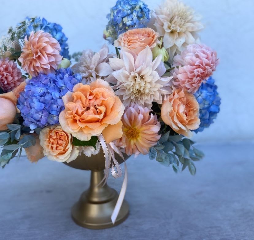 Golden Coast Reverie  - A floral dream inspired by Southern California’s sunlit horizon — where ocean breeze meets golden light. Soft peach garden roses with apricot and blush dahlias drift alongside sky-blue hydrangeas, capturing the delicate harmony of coast and sky. Designed in a brushed gold compote, this piece evokes the warmth of sunset and the calm of sea air — a reverie of elegance, nostalgia, and quiet luxury that embodies coastal California living. 