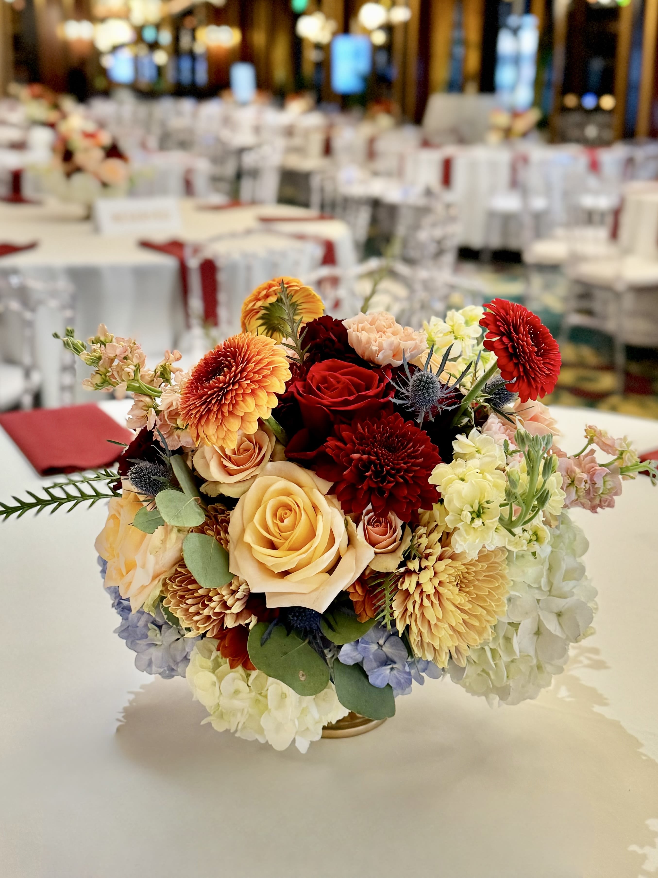 Table Centerpieces - A beautiful table centerpiece can transform an ordinary meal into an extraordinary experience. Imagine an elegant table setting, its warm tones contrasting with the soft glow of candlelight. At the center, a lush arrangement of flowers in soft, harmonious hues — pale blush carnations, red roses, and deep burgundy daisies — are nestled among sprigs of fresh greenery like eucalyptus and fern. The flowers are artfully placed in a low, elegant gold compote, allowing conversation to flow easily across the table. Imagine yourself being the best host that your guests will talk about for days, weeks, even years to come.   Flowers will be in season. Perfect for holidays, thanksgiving, birthdays, celebrations, and private parties. 