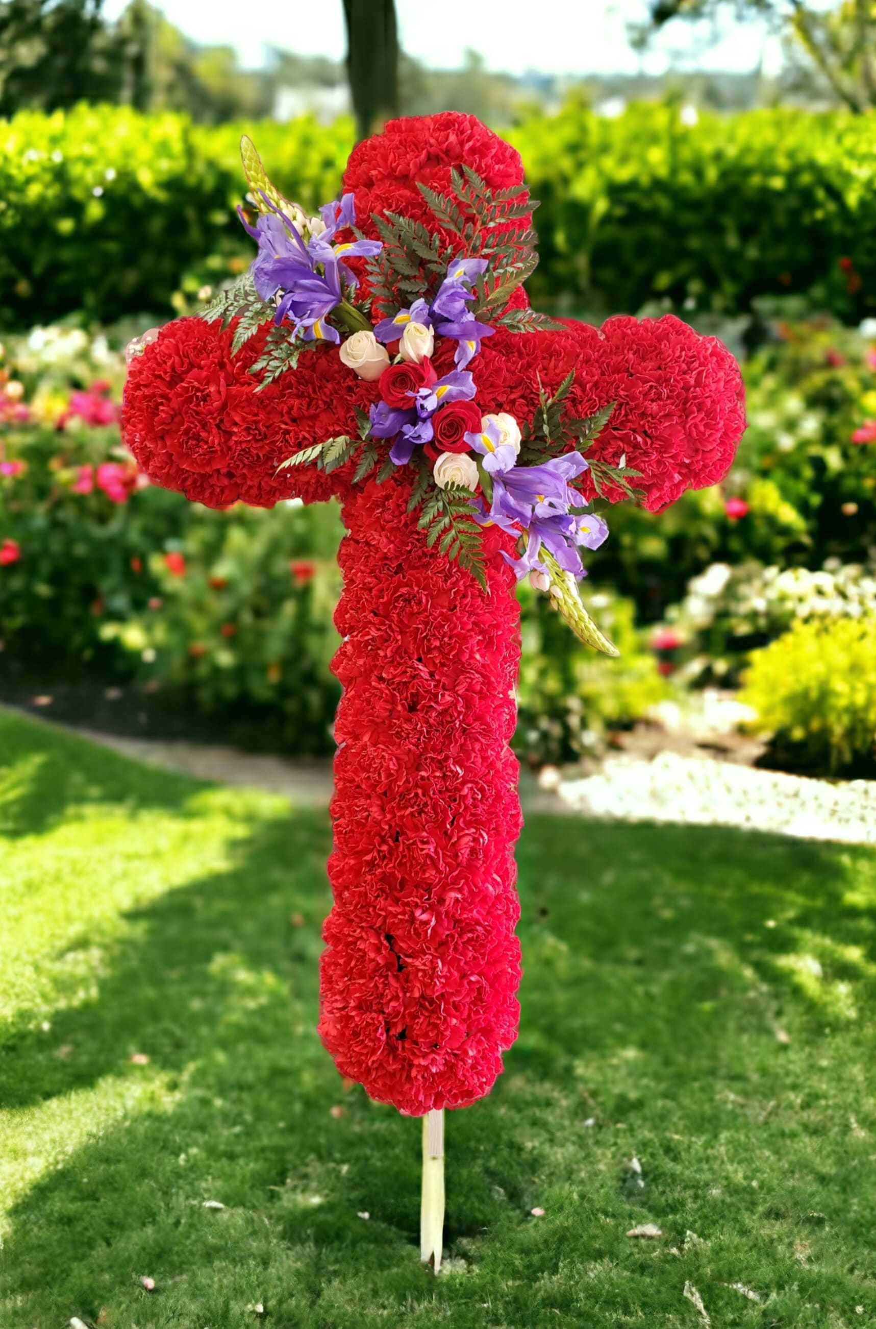  Faithful Hero Floral Cross - This thoughtful and meaningful tribute is a vibrant expression of faith. Designed by one of our florist of bright red carnations, blue iris, roses and other complementary greenery makes a powerful statement that’s sure to be remembered and appreciated at a wake, funeral or graveside burial service. It comes with a metal easel for a floor/ground-standing display. Measures approximately 24"H x 18"W.