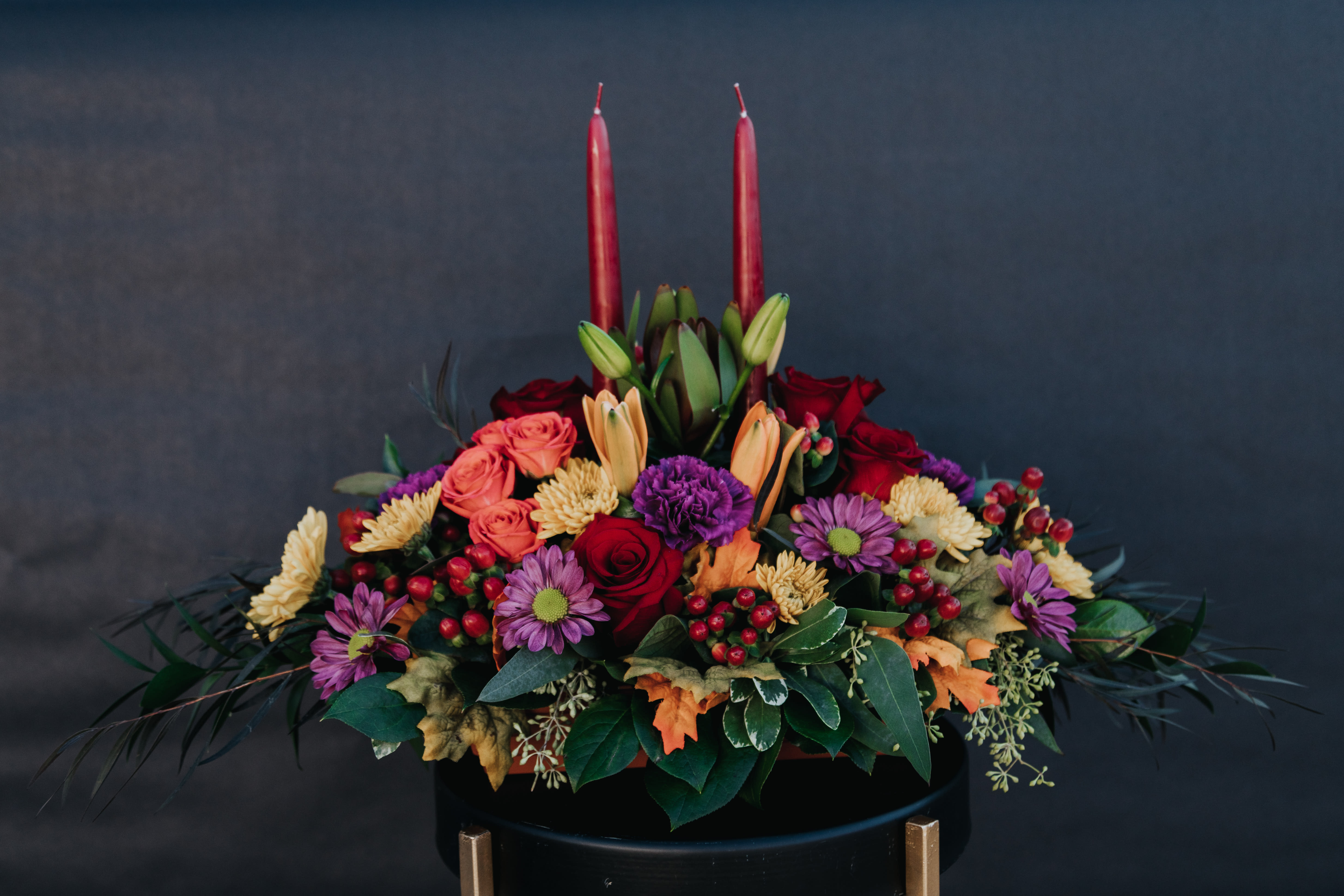 Autumn Jewel Centerpiece - A jewel toned arrangement of fall flowers just right for your holiday table.  Includes a bountiful assortment of blooms including red roses, coral spray roses, purple carnations, purple and fall daisies and freshly fallen autumn leaves.  Includes 2 burgundy taper candles.