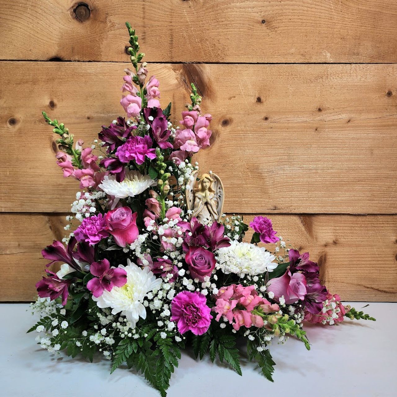 Angels Among Us - Step into a realm of grace and serenity with &quot;Angels Among Us,&quot; a flower arrangement designed to uplift and inspire. This enchanting display features a harmonious blend of flowers that evoke both elegance and tranquility.  At the heart of this arrangement is a cherished keepsake angel statue, serving as a divine focal point. Surrounding this celestial figure is a soft and soothing palette of blooms that add a touch of purity and grace, their subtle elegance perfectly complementing the statue's presence.  Together, these elements create a beautiful tribute that celebrates the divine and the delicate, making &quot;Angels Among Us&quot; a perfect centerpiece for honoring loved ones or bringing a touch of ethereal beauty into any space.  approx 21&quot; H x 21&quot; W  PLEASE NOTE: Some flowers, vases and other products pictured are subject to availability, and may be substituted for a similar item if unavailable according to our substitution policy.
