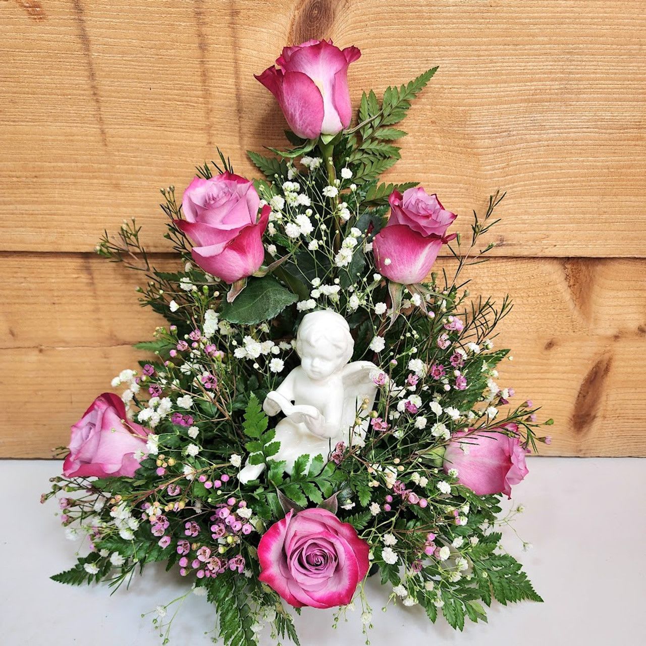 Love From Above - &quot;Love from Above&quot; is a serene and heartfelt floral arrangement designed to evoke peace, love, and heavenly beauty. At the heart of the display sits a delicate porcelain cherub figurine, symbolizing purity, grace, and the comforting presence of a guardian angel.  Surrounding the cherub are vibrant pink roses, representing love, admiration, and appreciation. Interwoven among the roses are soft sprays of white baby's breath and small pink waxflowers, adding lightness and texture to the arrangement. Lush green ferns and foliage form a natural base and backdrop, enhancing the vibrant colors and giving the piece a fresh, organic feel.  Perfect as a sympathy tribute, a spiritual gift, or a centerpiece to honor a loved one, &quot;Love from Above&quot; serves as a gentle reminder of everlasting love and the beauty of celestial guidance.  approx. 14&quot; H x 12&quot; W  PLEASE NOTE: Some flowers, vases and other products pictured are subject to availability, and may be substituted for a similar item if unavailable according to our substitution policy.