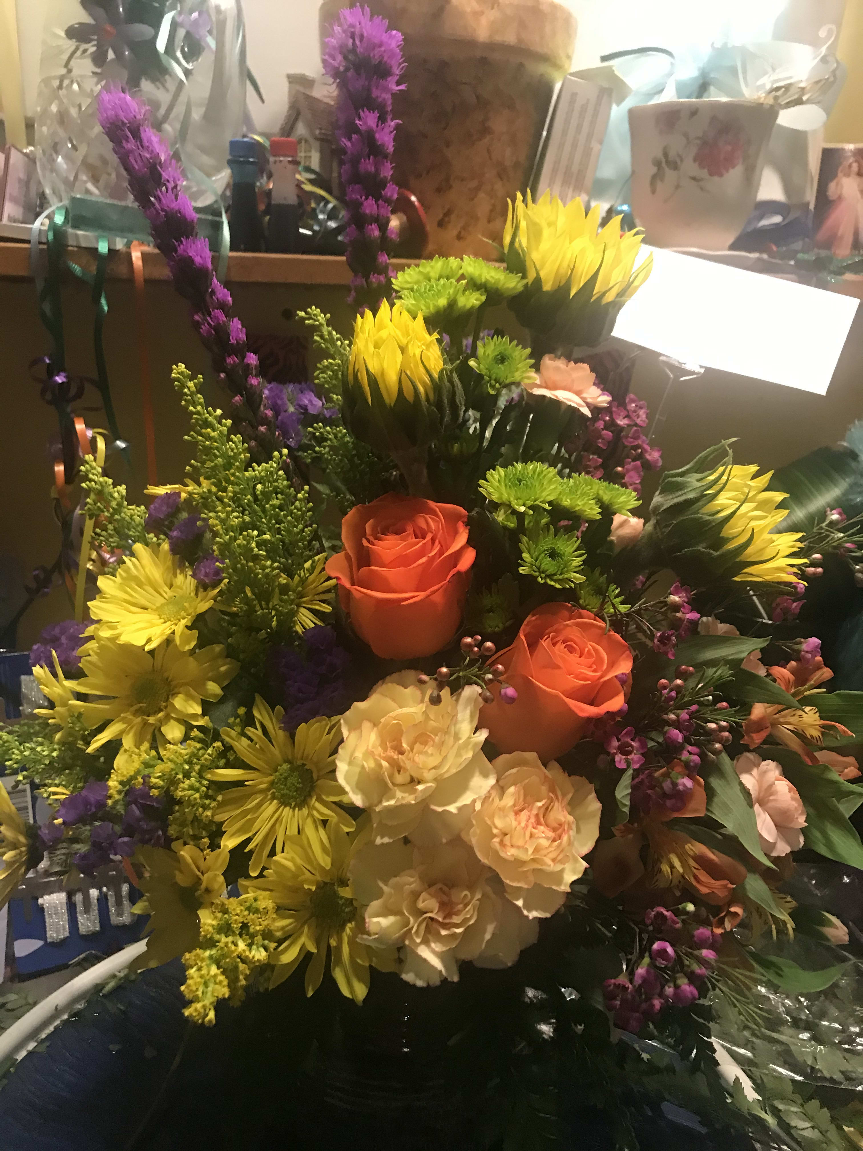 Sun Soaked Blooms - As beautiful as a summers garden - this bright bouquet shows sunflowers reaching toward the sun surrounded by roses, daisies, liatris and alstromeria in one of our many containers