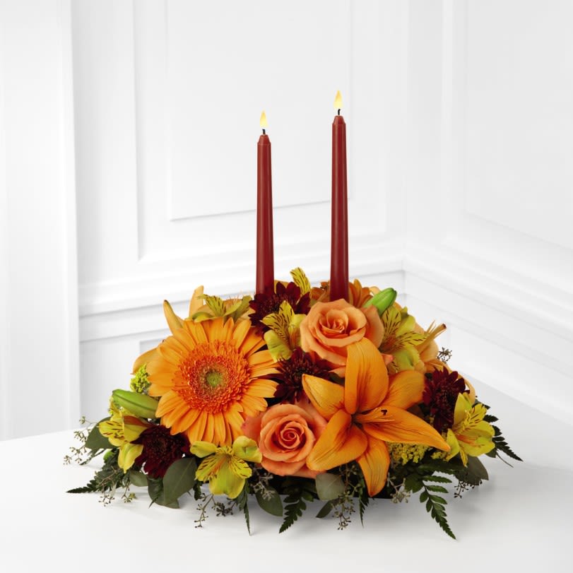 The FTD Bright Autumn Centerpiece - The FTD Bright Autumn Centerpiece spins the magic of the Fall season with each sun-kissed petal to set your gathering space aglow with blooming beauty.  Two taper candles are surrounded by a gorgeous arrangement consisting of orange Asiatic Lilies, gerbera daisies, and roses with golden Peruvian Lilies, burgundy cushion poms and assorted greens to create the perfect addition to your Autumn celebration.  