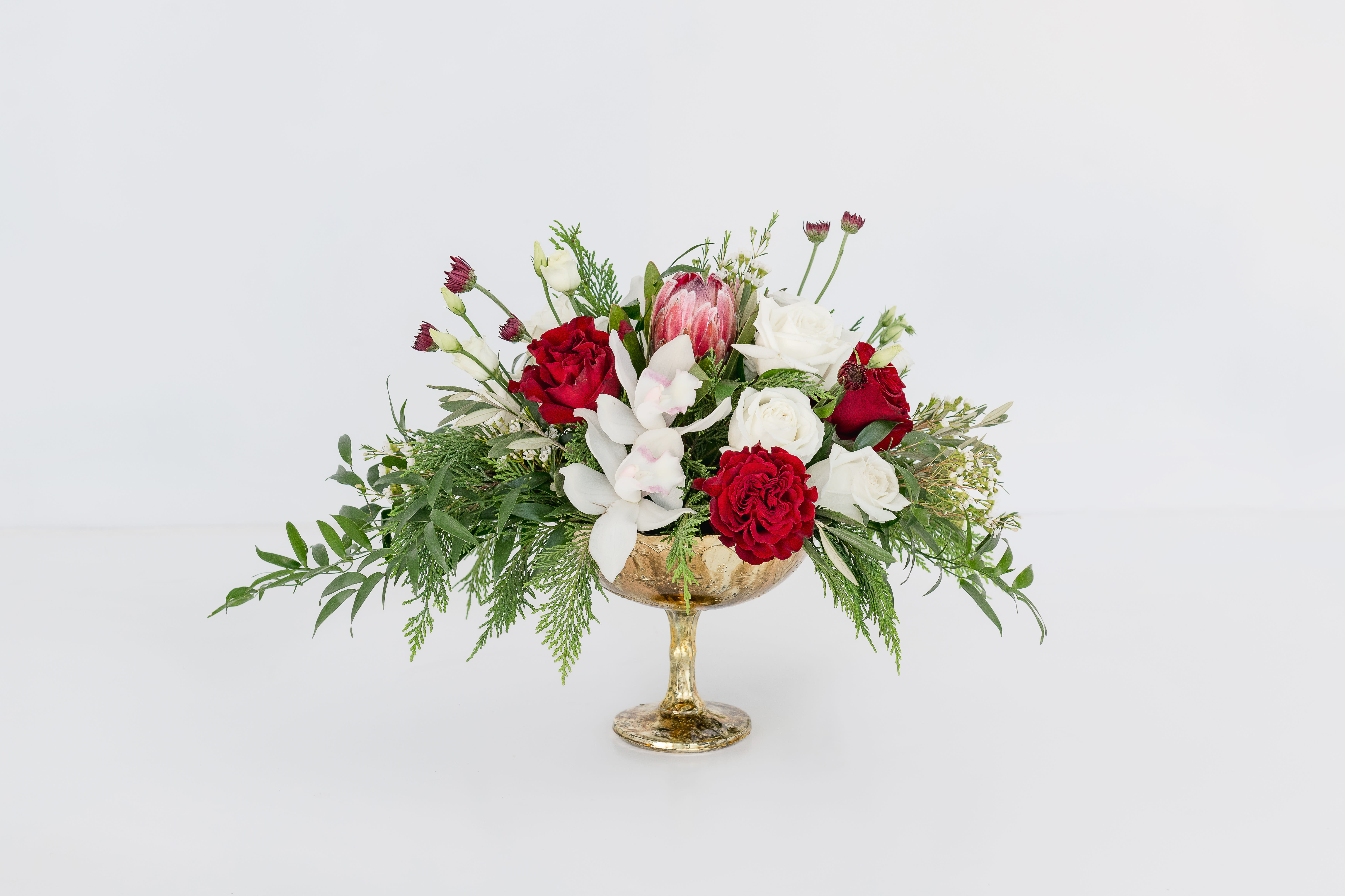 Joyful Collection Compote Arrangement  - Breathtakingly beautiful -- and full of seasonal charm. A beautiful mercury glass compote style container is overflowing with a timeless blend of red, white, and holiday greens. This design showcases a lush arrangement of orchids, roses, mums, berries, King Protea, and festive greenery—breathtakingly beautiful and full of seasonal charm. This elegant design makes the perfect centerpiece for any holiday celebration, or a lovely gift or a décor piece for home or office.  Product Details: This arrangement is approximately 14&quot; tall and 10&quot; wide. Upgrade to the Premium size for an additional 10 stems of flowers and greenery. 