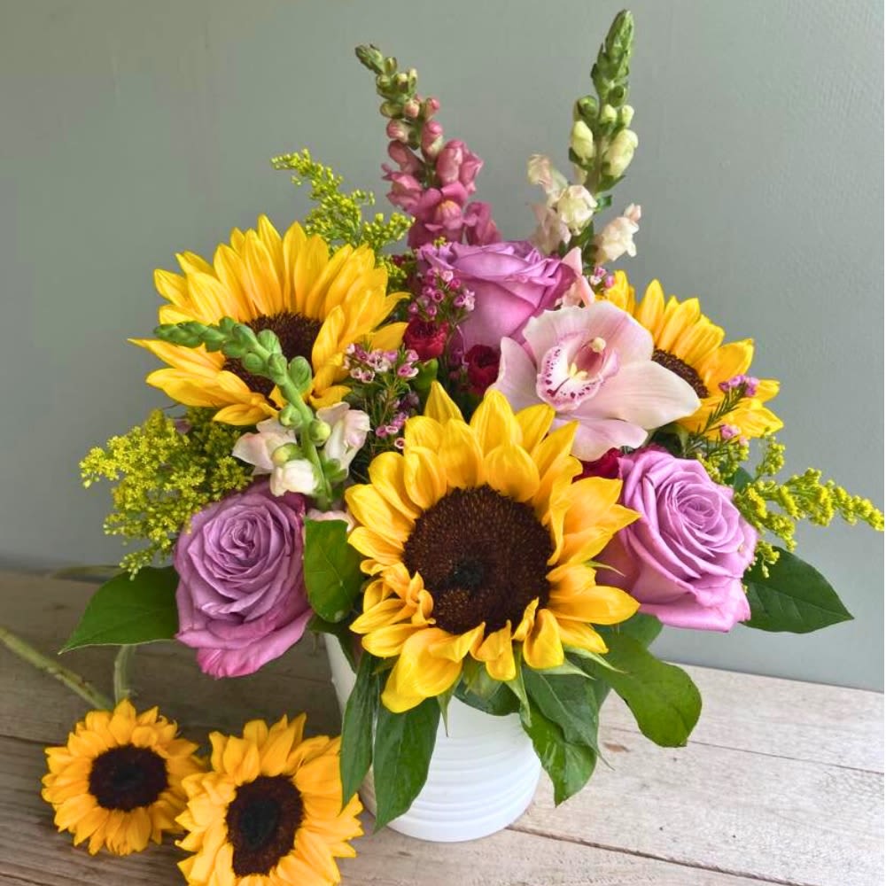 Sunny Smiles  - This stunning arrangement features a vibrant mix of sunflowers and purple roses elegantly displayed in a vase. Perfect for brightening up any room, this bouquet exudes a sense of warmth and cheerfulness. Ideal for gifting on special occasions or simply as a beautiful addition to your own home decor, this floral masterpiece is sure to make a lasting impression.   A unique arrangement hand created by our experienced designers. Please note that every design is custom made and substitution to container and flower types may happen from time to time. Design aspects will always stay the same, but if needed, we will substitute with flowers of equal or greater value to offer you a great floral shopping experience! Our flowers are 100% guaranteed. Please call us with any questions or concerns.