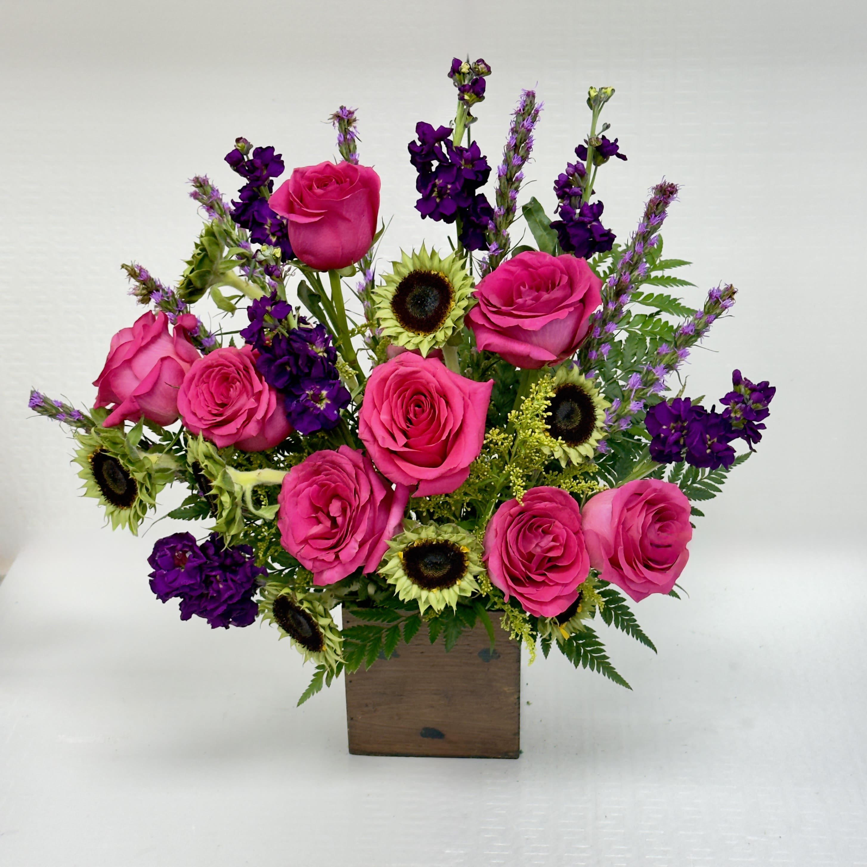 Fuchsia Sunrise - A vibrant mix of hot-pink roses, deep purple blooms, and mini sunflowers arranged in a rustic wooden box- perfecr for brightening any space withbold, cheerful color.