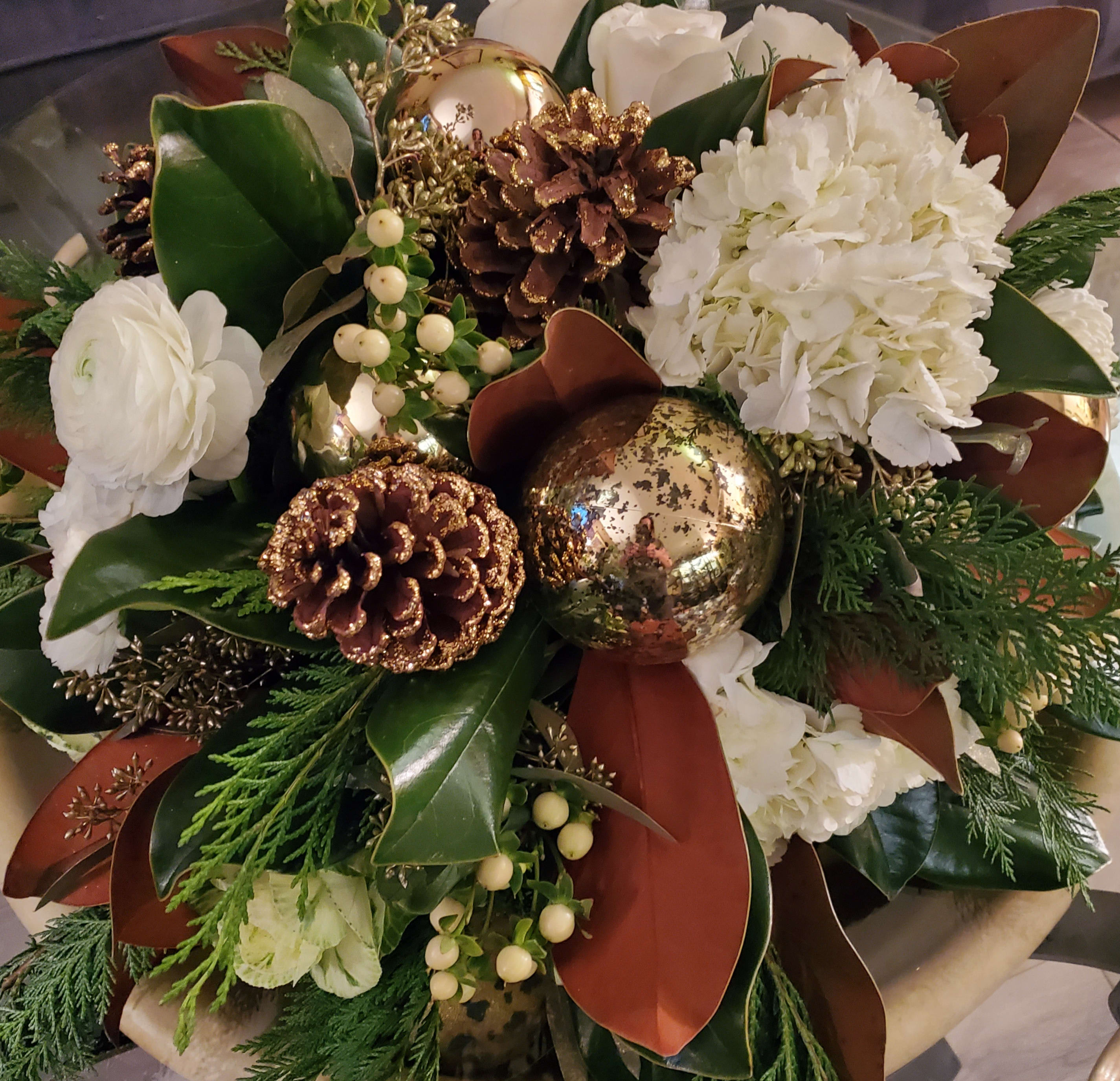 Joan of Arc - Consisting of Magnolia, Seeded Eucalpytus, Roses, Hydrangeas, Ranunculus, Hypericum Berries, Cedar, Gold Glitter Dipped Pinecones, and Christmas Ornaments, this is a posh take on the classic Christmas floral arrangement. This show-stopping winter bouquet will make your home cozy and festive. 
