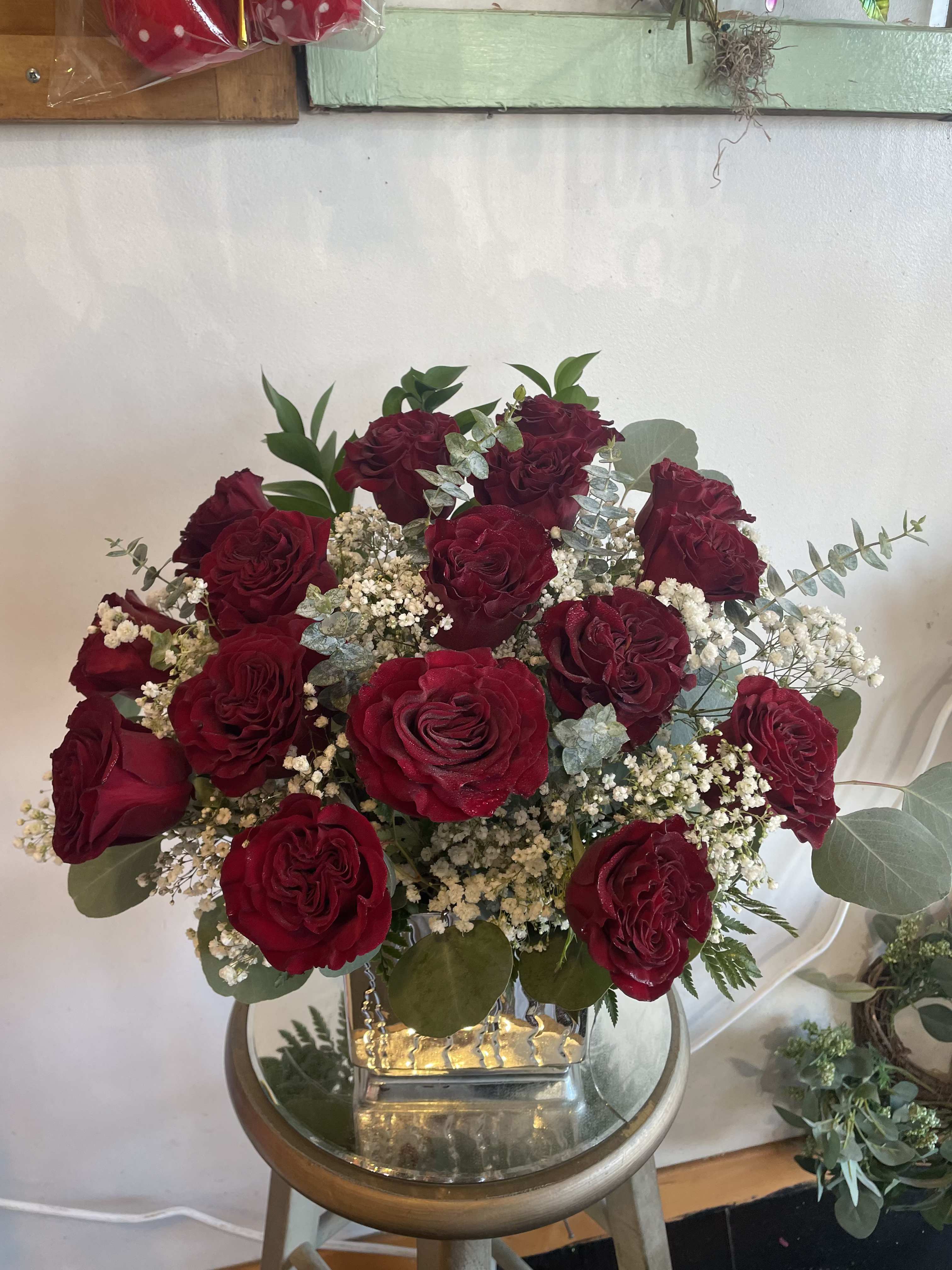 Velvet Romance Arrangement - A stunning floral arrangement featuring deep heart red roses elegantly displayed in a square, reflective silver or gold  vase. The roses are full and velvety, surrounded by delicate white baby's breath and accented with soft eucalyptus leaves and lush greenery.  creating a warm and inviting presentation. This arrangement exudes romance, sophistication, and timeless beauty.