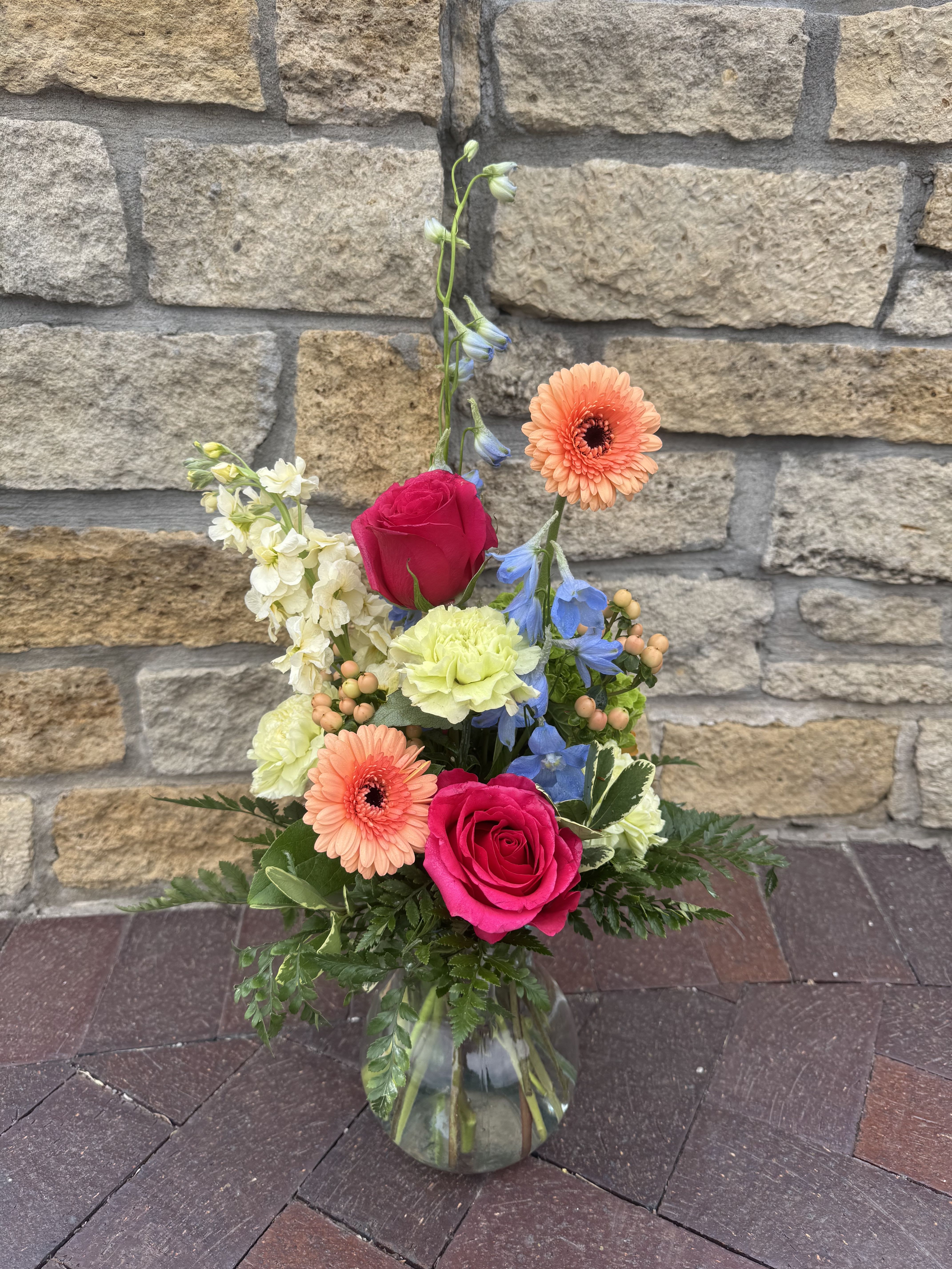Forever Julia - This arrangement features fresh roses, mini gerbera daisies, vibrant blue delphinium, carnations, hypericum berries, and finished with fragrant stock.