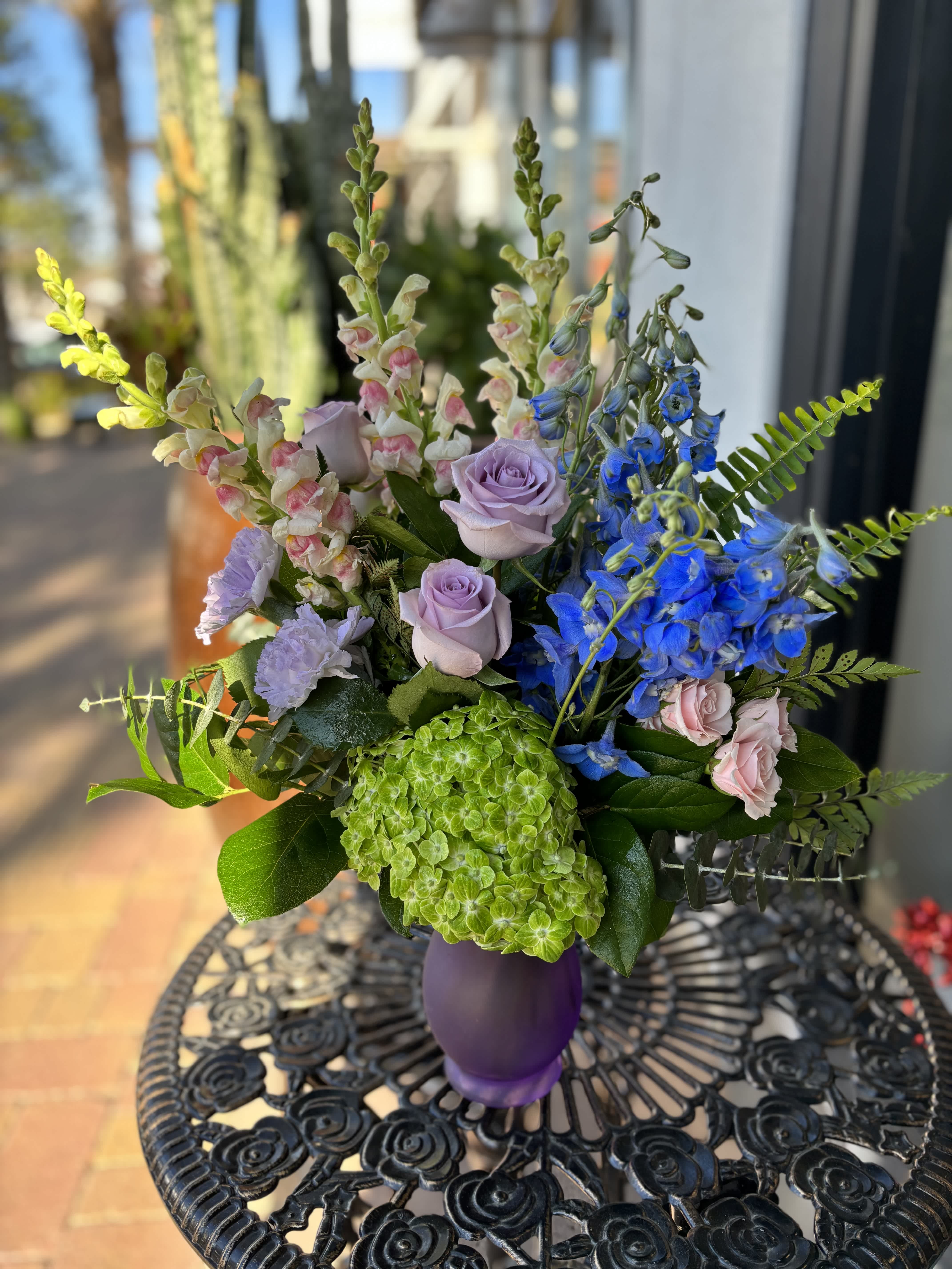 Candy Sky - Pink Snapdragons, light blue delphinium, lavender roses, lavender carnations, hydrangea, and pink spray rose with mixed foliage arranged in a purple glass vase.  