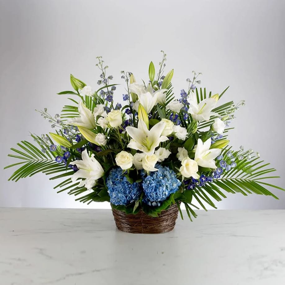Deepest Condolences - A blue, white, and green funeral basket for any type of service. Held in a wicker basket, large palms spread out and give a natural spotlight for the feature blooms and colors.  Approximately 28” W x 20” H