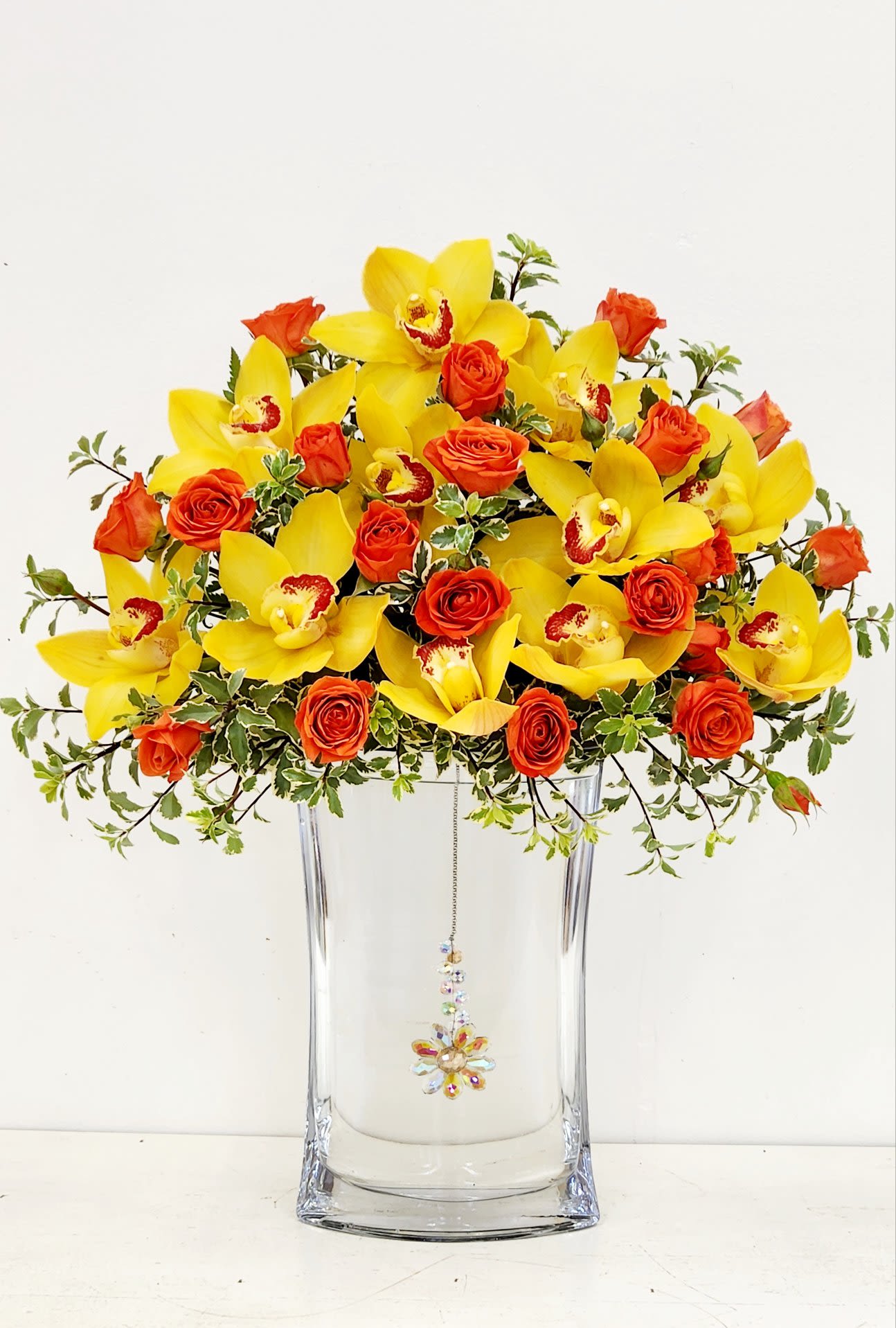 Whispers of Elegance - A stunning display of yellow orchids and orange spray roses, artfully floating atop a tall clear glass vase. A delicate pendant suspended below adds a graceful sparkle, creating a design that feels both radiant and refined — a true whisper of elegance and warmth.  PREMIUM SIZE ONLY 20&quot;H by 16&quot;W