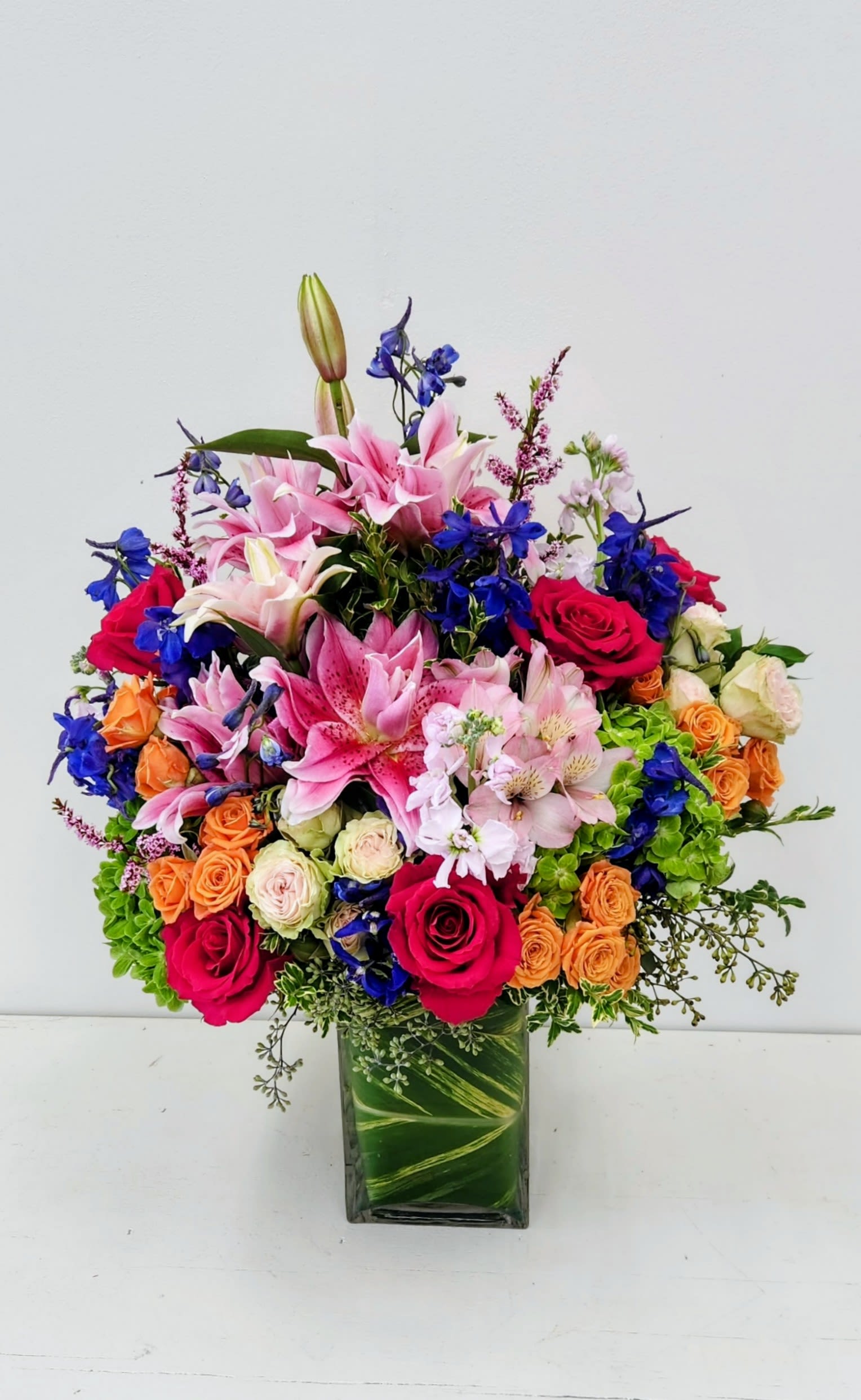 You're in My Thoughts Bouquet - An exquisite floral arrangement showcasing a harmonious blend of elegance and luxury. At its heart, soft pink rose lilies exude grace, accompanied by vibrant orange spray roses that add a splash of warmth. Lush green hydrangeas provide a rich, textural contrast, while stunning blue delphiniums introduce a captivating pop of color. This breathtaking composition is perfect for elevating any space with its sophisticated charm and enchanting beauty.