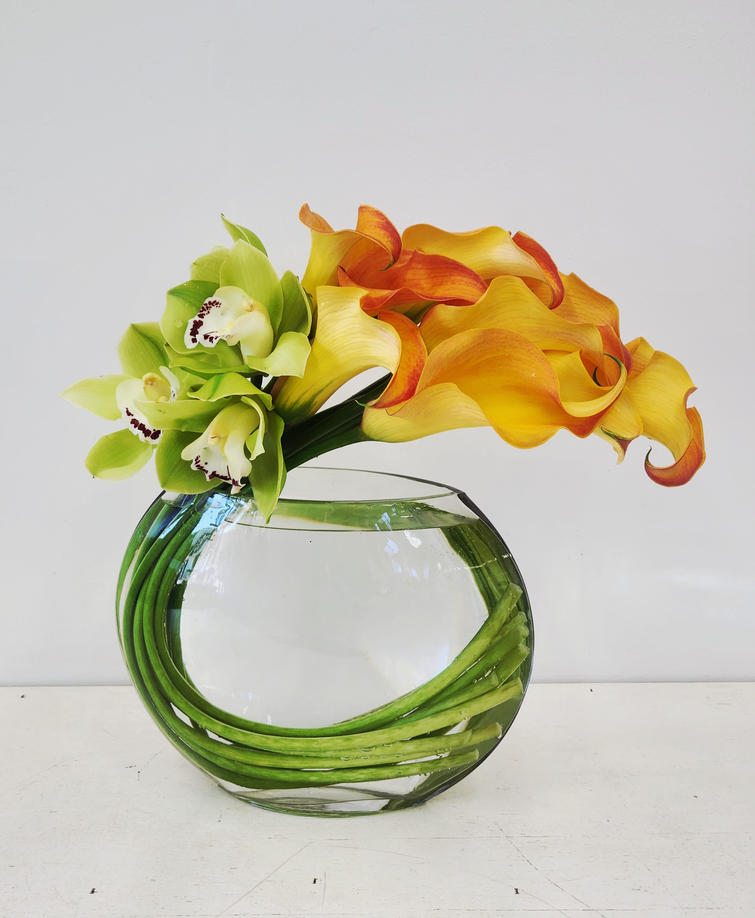 Sensational - Striking orange calla lilies arranged in a perfect circle create an eye-catching focal point, nestled in a sleek circular vase. Lush green orchids accent the composition, adding depth and sophisticated contrast. This modern arrangement brings warmth and elegance to any space, making it an ideal choice for celebrations, apologies, or simply brightening someone's day.  PREMIUM SIZE ONLY 15&quot; BY 15&quot;