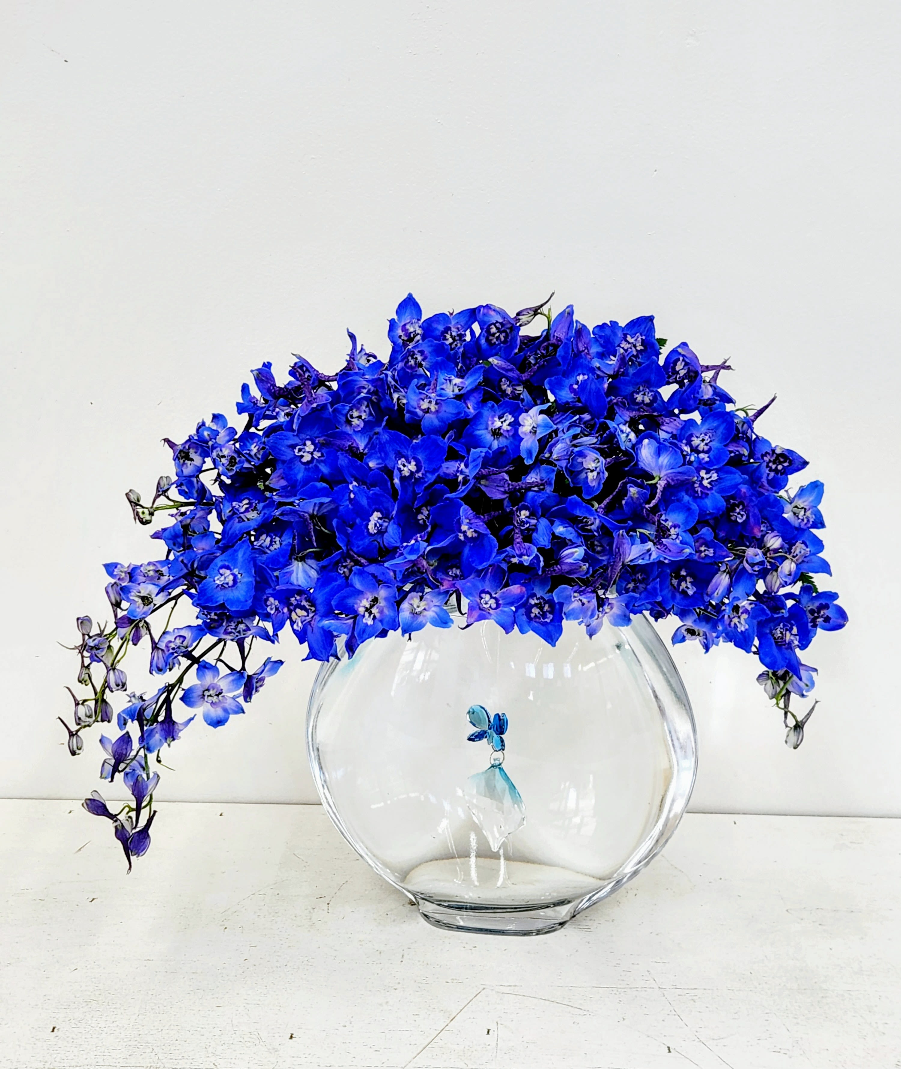Blue Magic - Blue Magic presents an exquisite fusion of botanical artistry and contemporary design. Voluminous blue delphiniums cascade gracefully above a refined circular vessel, while an elegant blue pendant suspended within creates an intriguing visual narrative. The inverted half-moon silhouette of blooms paired with the pendant's subtle presence elevates this arrangement beyond traditional floristry—a curated statement piece for discerning collectors who appreciate understated luxury and thoughtful craftsmanship.  ONE SIZE ONLY: PREMIUM 15&quot; by 18&quot;