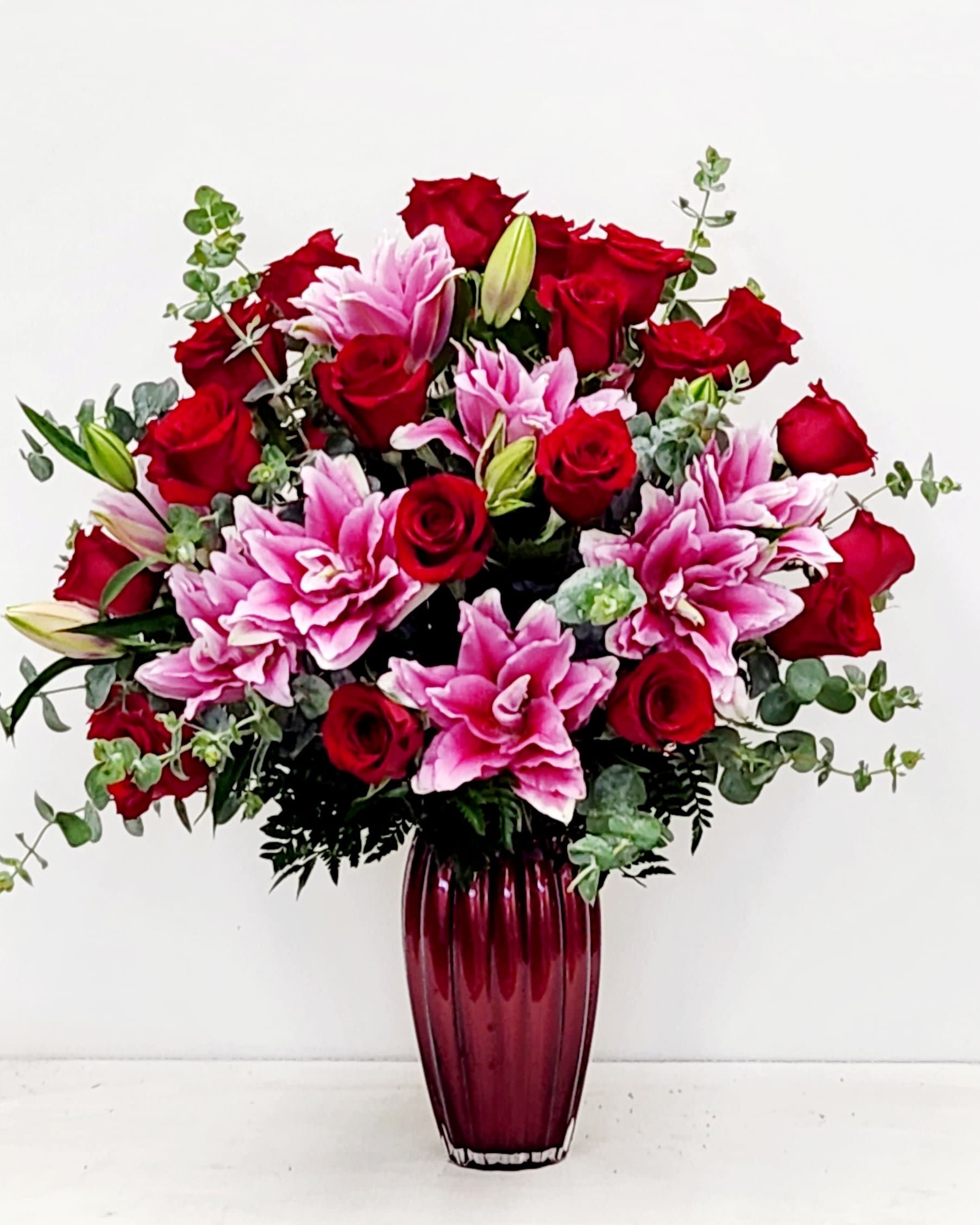 Love Is Forever - A masterwork of floral design featuring premium red roses and blush pink lilies, artfully composed with graceful spiral eucalyptus. Presented in a striking crimson designer vessel, this arrangement embodies understated elegance and timeless romance. Each element is carefully selected to create visual harmony and depth. An exceptional choice for those who appreciate refined beauty and meaningful gestures.  ONE SIZE ONLY DESIGNED WITH 18 PREMIUM RED ROSES. 18&quot; WIDE BY 22&quot; TALL