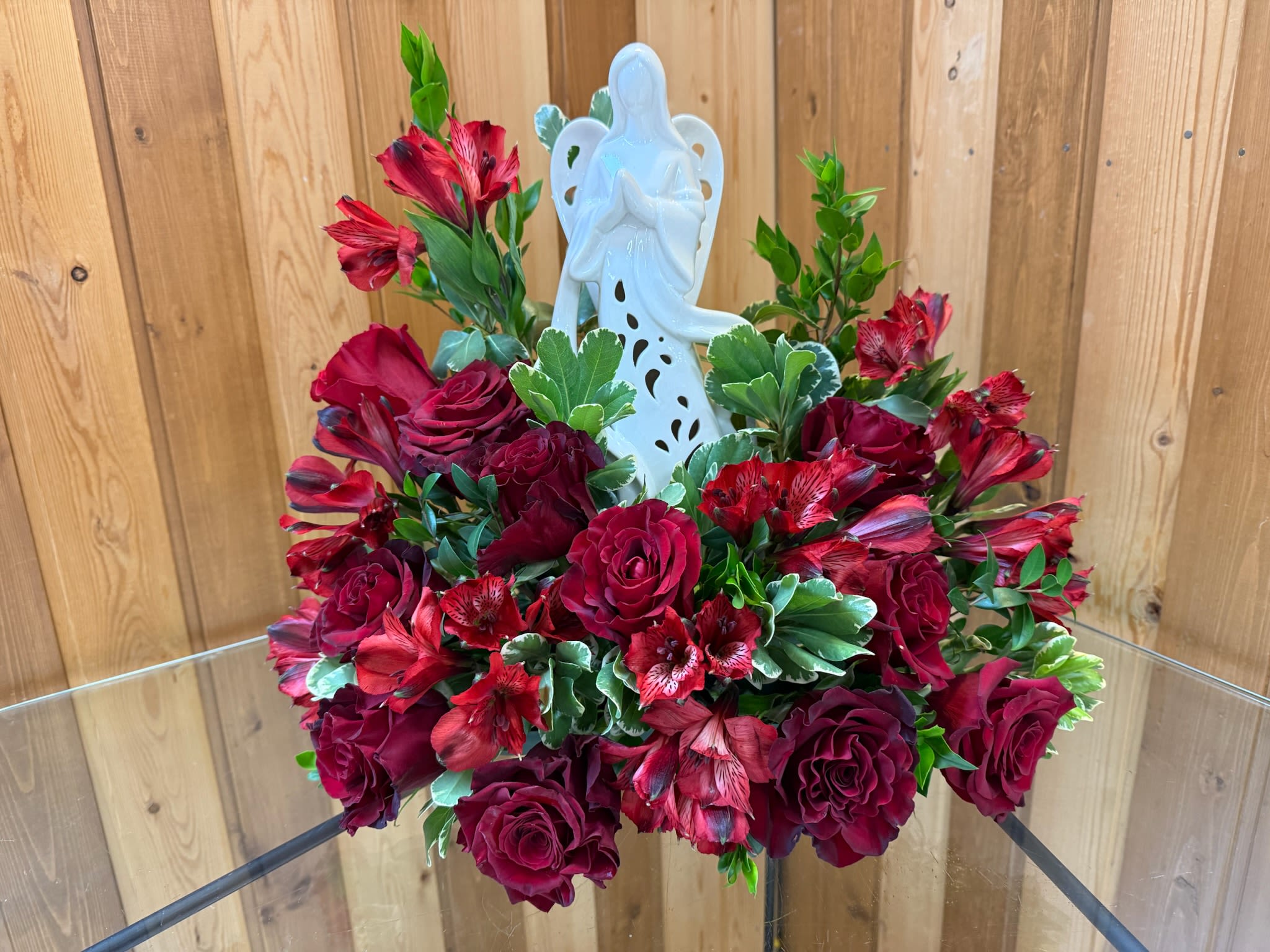 Eternal Embrace Angel Tribute - This graceful and heartfelt arrangement is a symbol of love, protection, and remembrance. A serene white angel figurine stands at the center, embraced by a stunning display of deep red roses, representing love, devotion, and cherished memories. Accented with soft greenery and hints of eucalyptus, this arrangement exudes elegance and peace. ***ANGEL MAY VARY BUT WILL BE SIMILAR  A meaningful sympathy tribute, perfect for honoring a loved one’s memory and offering comfort to those who hold them close in their hearts.