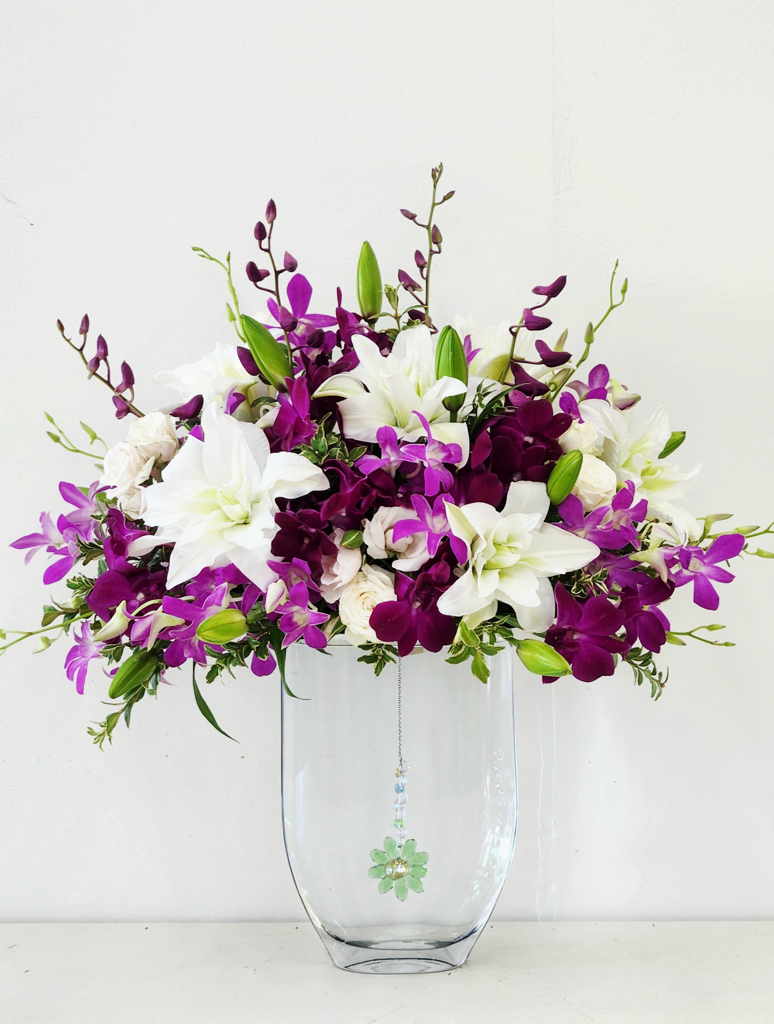 You Inspire Me - Purple orchids and delicate white rose lilies dance around a stunning tall glass vase, creating an ethereal floating arrangement that catches the light beautifully. Soft green accents add dimension while a mysterious gem dangles inside the vase, sparkling beneath the blooms. This whimsical design brings a touch of magic to any space—perfect for inspiring someone special or treating yourself to a moment of floral joy. A piece of inspiring masterpiece. ONE SIZE ONLY. 18&quot;W by 22&quot;H