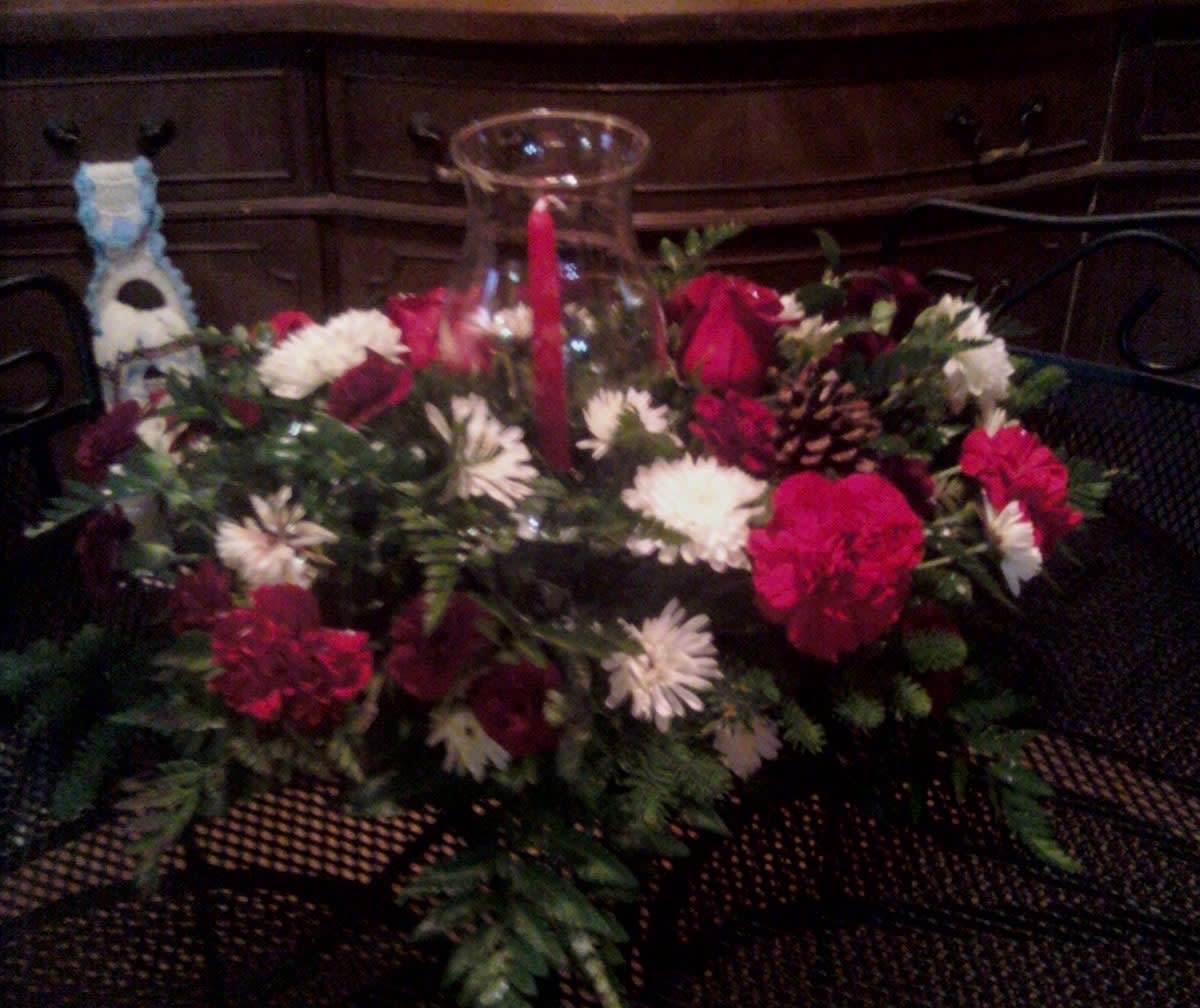 Dressed for the Season - Dressed for the season.  We can create your centerpiece to match your mood. Fall, Thanksgiving, Christmas or New Year's Eve. Let our designer's create a one-of-a-kind masterpiece that will WOW you and your guest's. Our standard size starts with greenery and mini carnations, regular carnations and seasonal flowers. Our deluxe adds in a few roses and seasonal accents. Our premium selection will showcase roses as the main blooms and seasonal flowers along with glitter sticks. All come with the glass hurricane in the middle of the design with one tapered candle. (Deluxe shown)