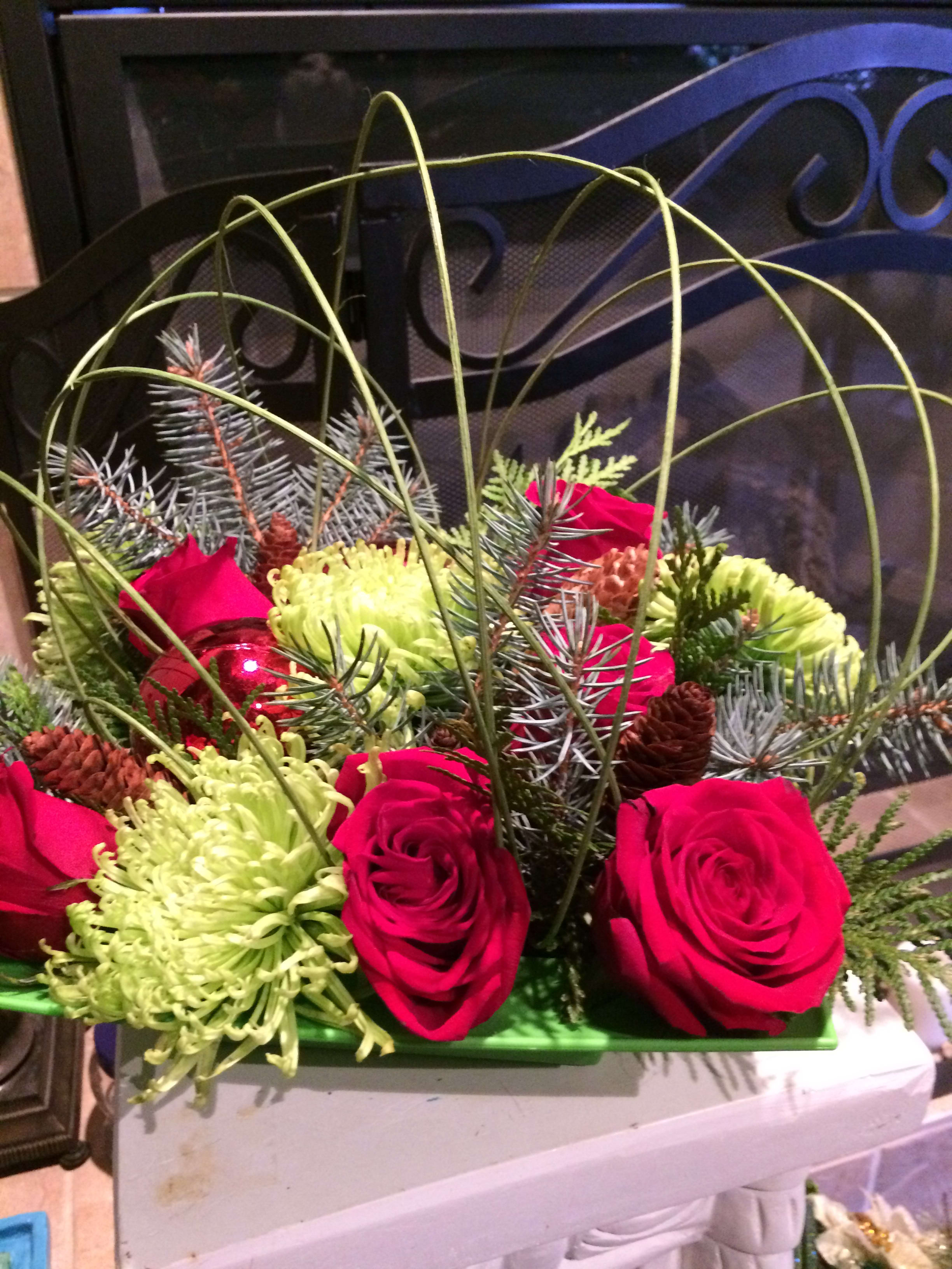 Crowning Glory Holiday Centerpiece - 2 Shiny Red Ornaments displayed with 6 Red Roses, 3 Fiji mums, 6 assorted pinecones with mixed greens. Highlighted with crossing  Apple Green midollino sticks for height &amp; emphasis placed in a apple green reusable square bowl.