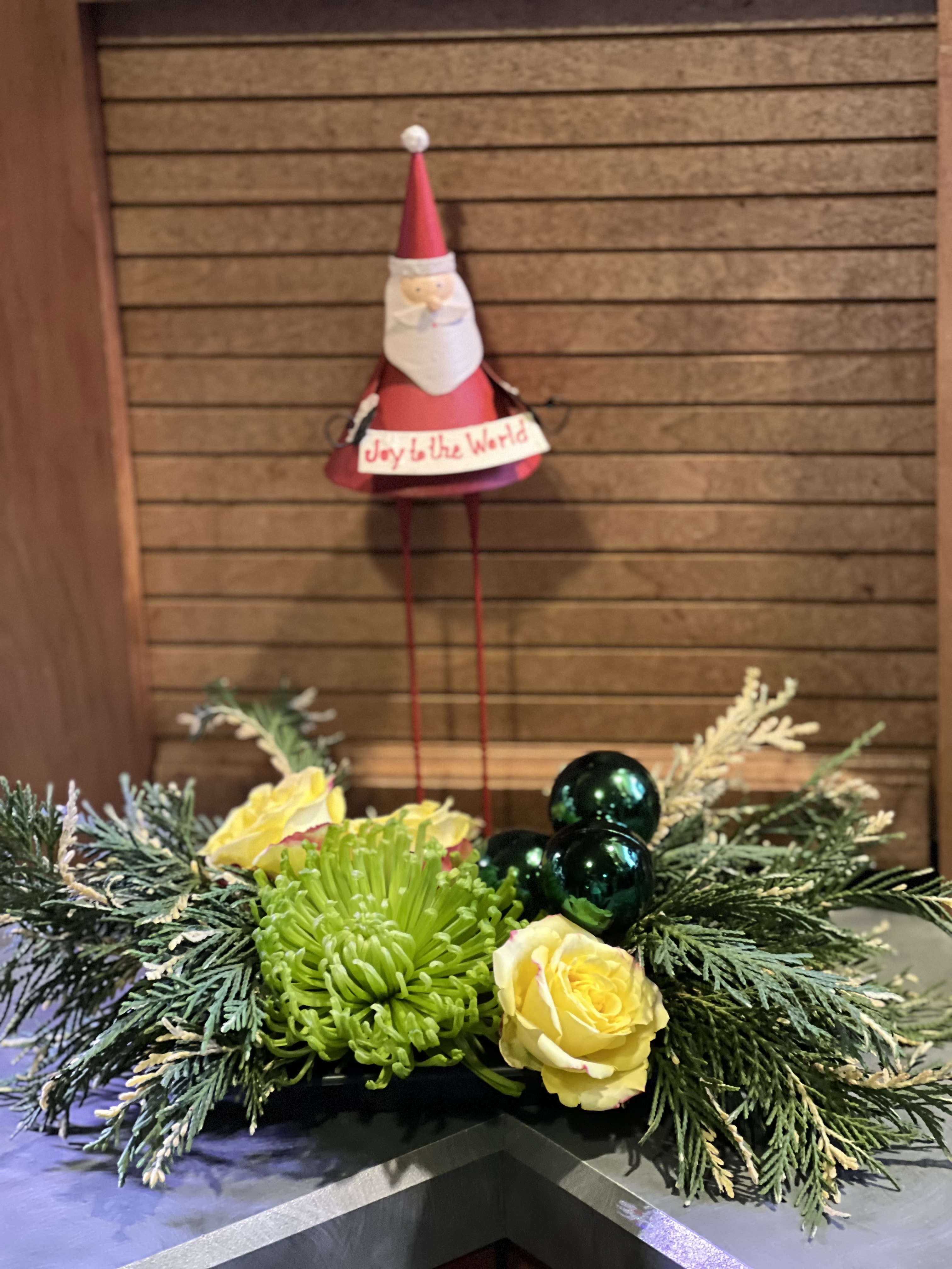 Santa and Scents - Santa keepsake with long legs paired with a tray of scented Holiday variegated greens. Accented with three sparkling ornaments. A Fiji chrysanthemum and a few spray roses. Perfect for the Holiday table or entrance end table. Approximate dimensions of Santa is 11&quot; tall. Tray with greenery and flowers is about 12&quot; wide.  The deluxe and premium will add in additional flowers.