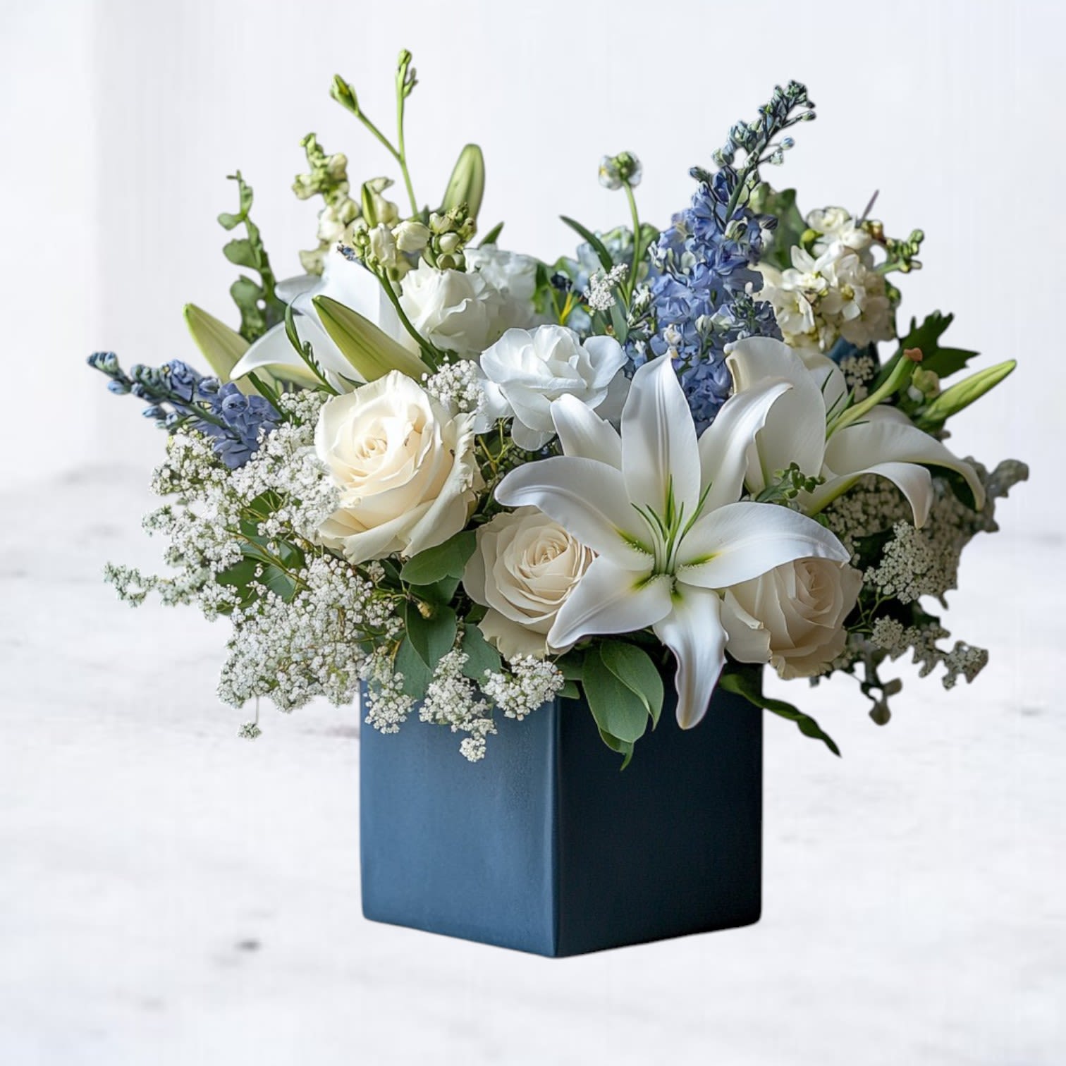 Midnight Tranquility - Bring a sense of serene beauty to a loved one’s memorial with our Midnight Tranquility floral arrangement. Designed in a deep navy cube vase, this peaceful composition features pristine white lilies, creamy roses, delicate baby’s breath, and soft blue delphinium, accented with fresh greenery. The soothing color palette offers a gentle, heartfelt expression of sympathy, perfect for honoring a life well-lived.  Whether delivered directly to a funeral home, memorial service, or a family’s home, this elegant sympathy arrangement conveys comfort and compassion when words alone aren’t enough.  Available for same-day delivery in Houston, Magnolia, The Woodlands, Conroe, Cypress, Spring, and surrounding areas. We also provide direct delivery to local funeral homes, including ensuring your tribute arrives beautifully and on time.