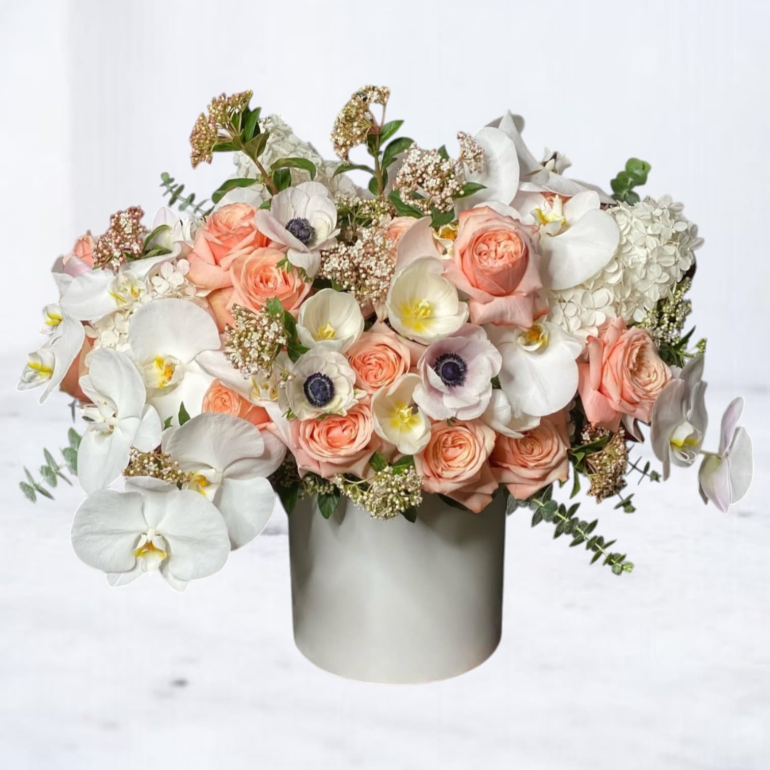 Halo Tribute Arrangement - Honor a life beautifully lived with our Halo Tribute Arrangement — an ethereal blend of soft peach roses, delicate white anemones, cascading white phalaenopsis orchids, and elegant touches of hydrangea and seasonal blooms. Designed in a modern ivory ceramic vessel, this premium sympathy arrangement conveys peace, grace, and everlasting love.  Perfect for funeral services, memorials, or a heartfelt condolence gift, Celestial Tribute offers a comforting expression of sympathy and remembrance. We offer same-day flower delivery in Houston and surrounding areas for funeral homes, churches, or private residences.