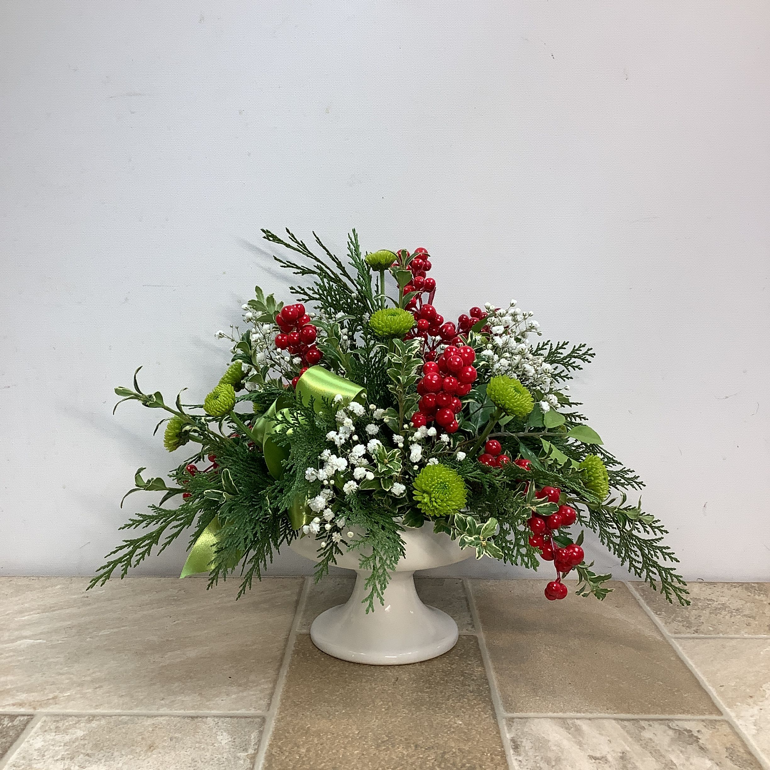 Evergreen Dream - Fresh evergreens, baby’s breath and bright green Kermit poms are expertly arranged in this classic holiday bouquet. The white pedestal vase and bright red faux berries complete this beautiful seasonal design. Approximate size: 14” wide x 12” tall.