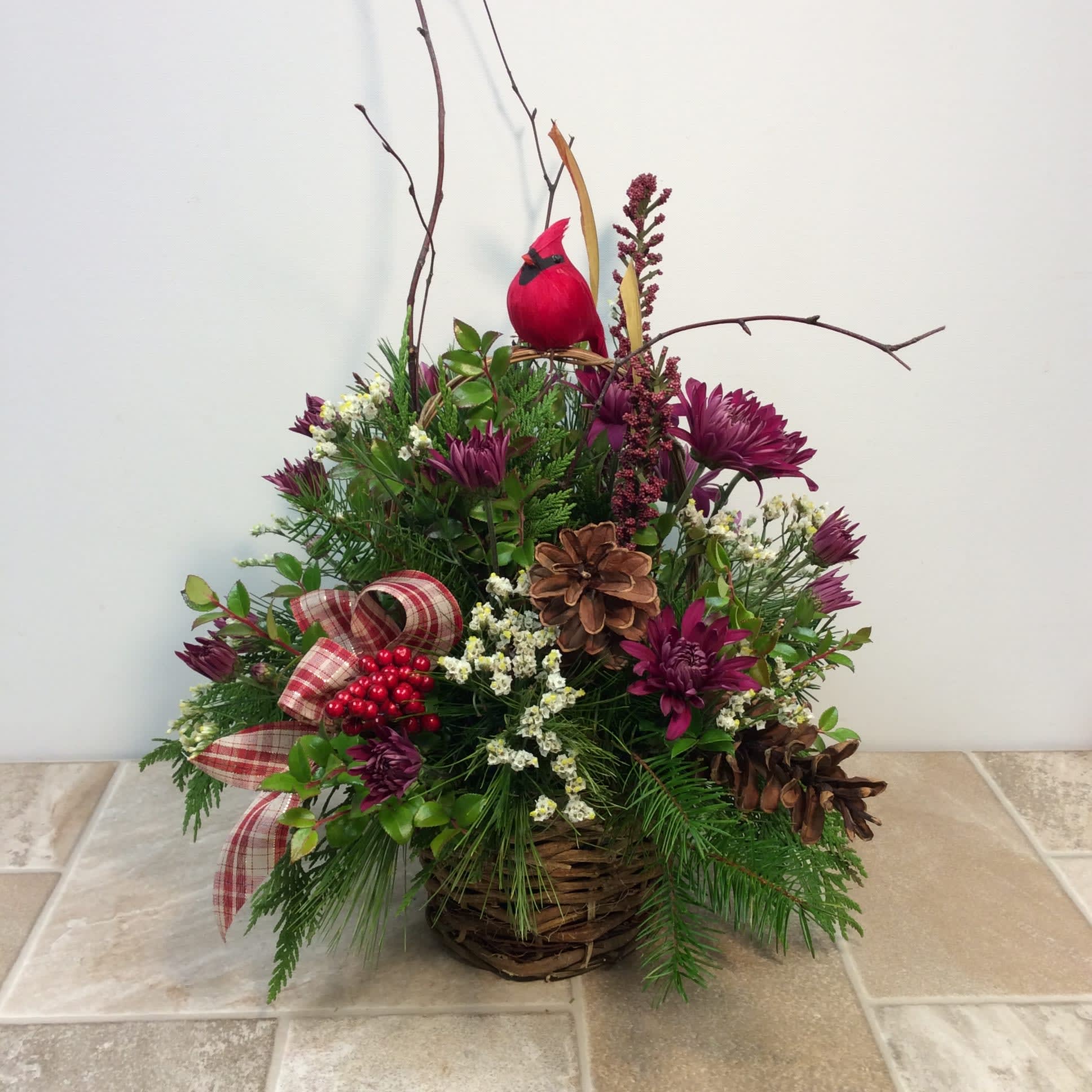 Clara - This basket arrangement is filled with mixed fresh evergreens, purple mums, red berries and white accent flowers. The twigs and pine cones help the cardinal perched on top feel right at home in this very long-lasting design.