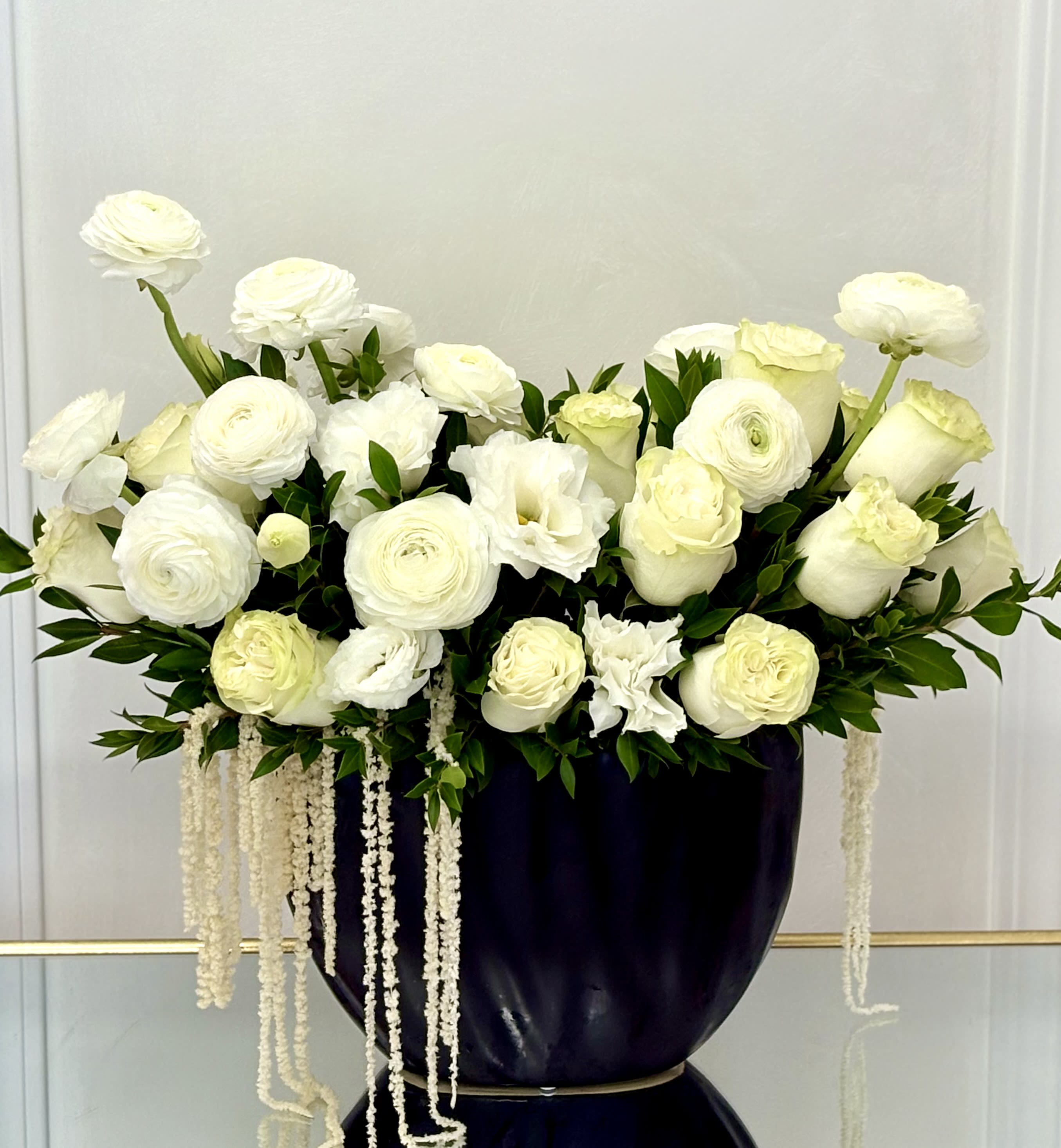 White Majesty By VLM Flowers - An exquisite display of white roses, ranunculus, and lisianthus with cascading amaranthus, arranged in a bold black vase. A stunning statement of purity and elegance, ideal for weddings, sympathy, or sophisticated décor.