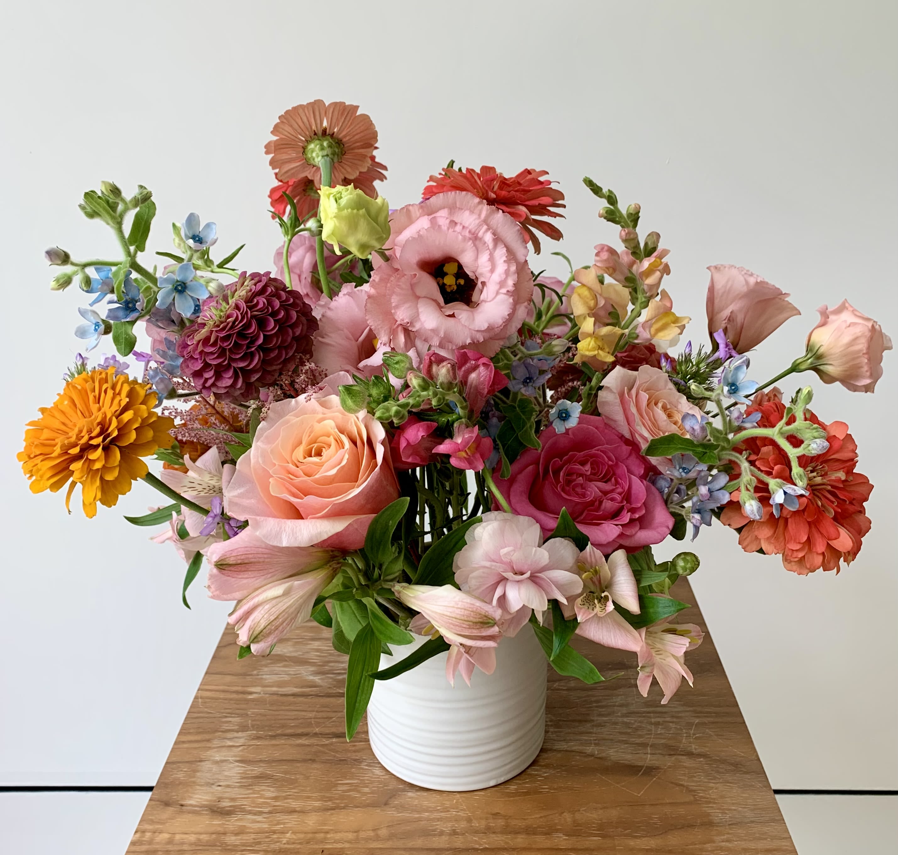 Floral Candy - A garden style flower arrangement in a white ceramic 5inch cylinder with focal flowers of snapdragons, tulips, hyacinths, roses, ranunculus, lisianthus, gerber daisies and alstromerias, with additional seasonal flowers and fillers to make a lovely candy like flower arrangement. The color palette may vary but there will always be some pinks, yellows, and blues.