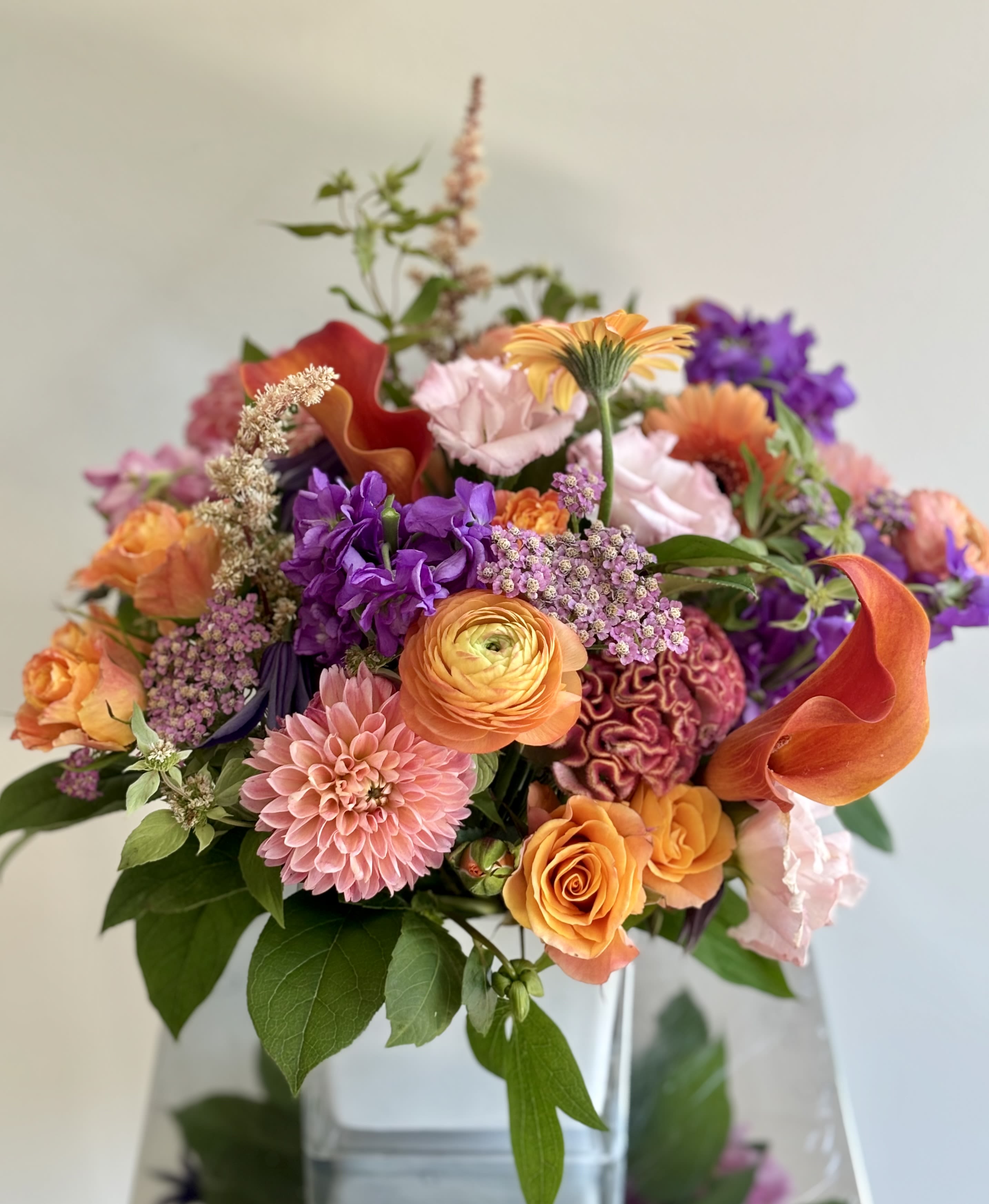 Fall In Love - Flower arrangement with beautiful fall premium flowers and floral goodies in orange-muted pink-purple color hues created in a glass cube vase. A mix of roses, ranunculus, dahlias, disbuds, callas, stock and more, embellished with green foliage. Colors may vary based on the availability.