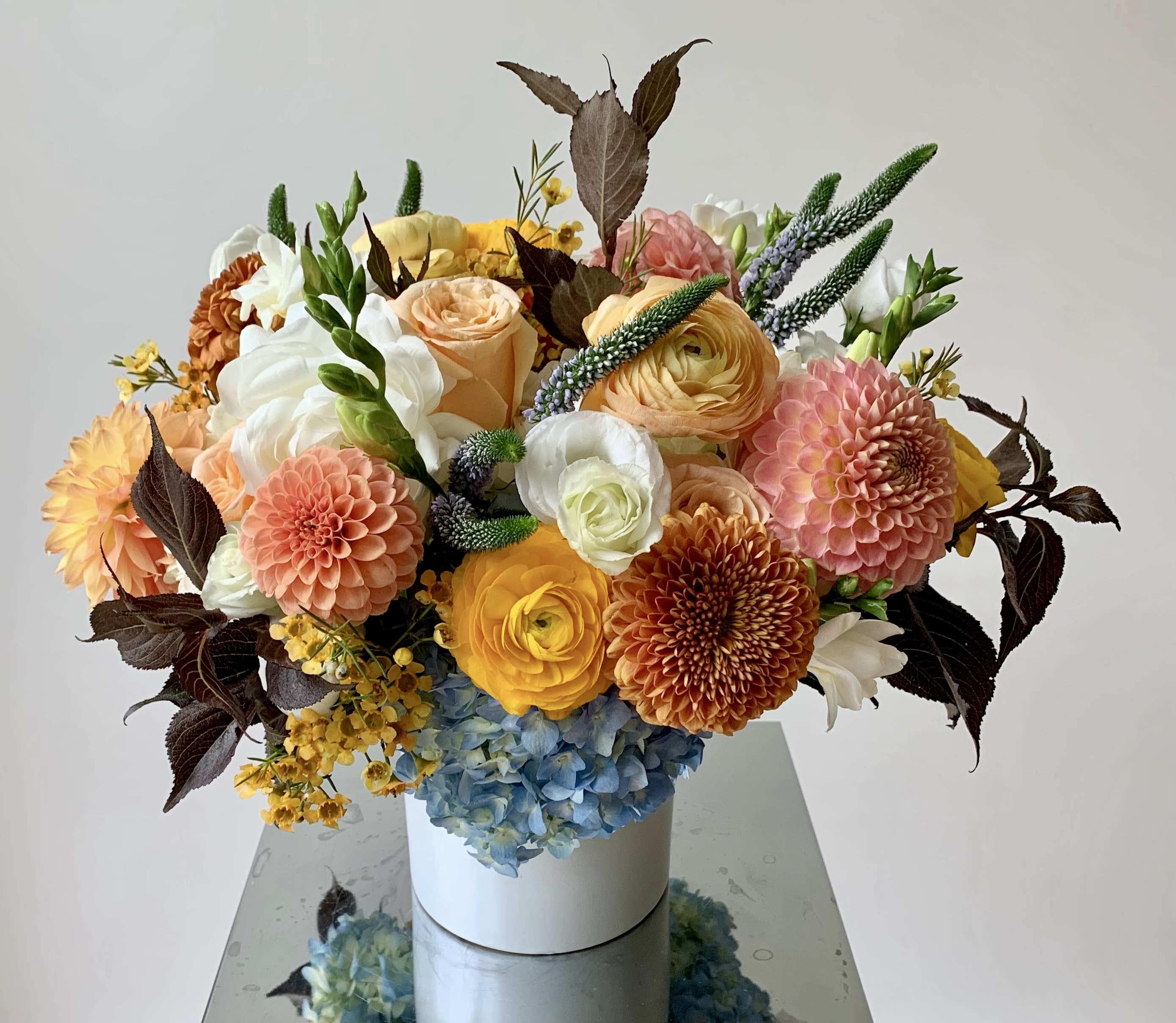 Soft Autumn - An autumnal flower arrangement of seasonal flowers in soft fall colors of yellows, orange, browns, bronze and whites nestled in light blue hydrangeas and fall foliage. The usual mix of flowers include roses, dahlias, ranunculus, cremons, lisianthus and more.