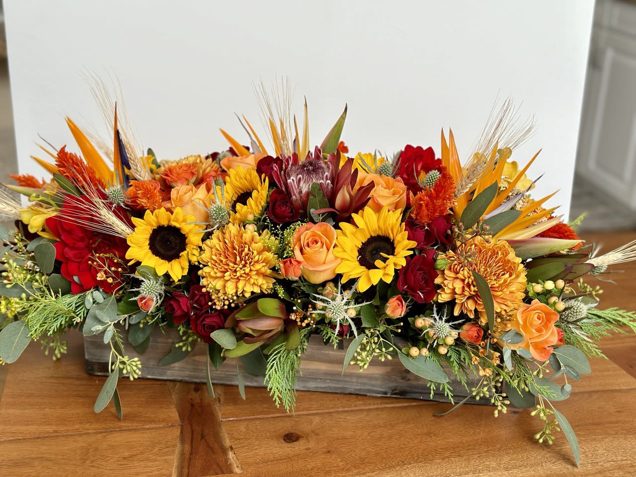 Warm Wishes Premium - As your loved ones gather around the table, they'll bask in the warm glow of fall flowers. Birds of paradise, red spray rose, red dahlia, sunflowers, orange rose, and fresh greenery delivered in rectangular wood. The wood container is 18.25&quot;x6.5&quot;. You can add up to 3 candles to this arrangement for FREE (please mention it in the Florist Instructions section).