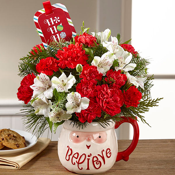 The Believe Mug Bouquet ( Christmas themed mug chosen by designer) - Sweet and stylish this holiday flower arrangement exudes the fresh and fun look  of Christmas joy in a Christmas mug you have come to know and love for years! Fanciful and oh so festive we bring together red roses carnations and mini carnations mingling with snowy white Peruvian lilies and an assortment of Christmas greens arranged to perfection in a keepsake ceramic Holiday mug . (mug will be not as shown for we have so many kinds. Designers choice based on size ordered