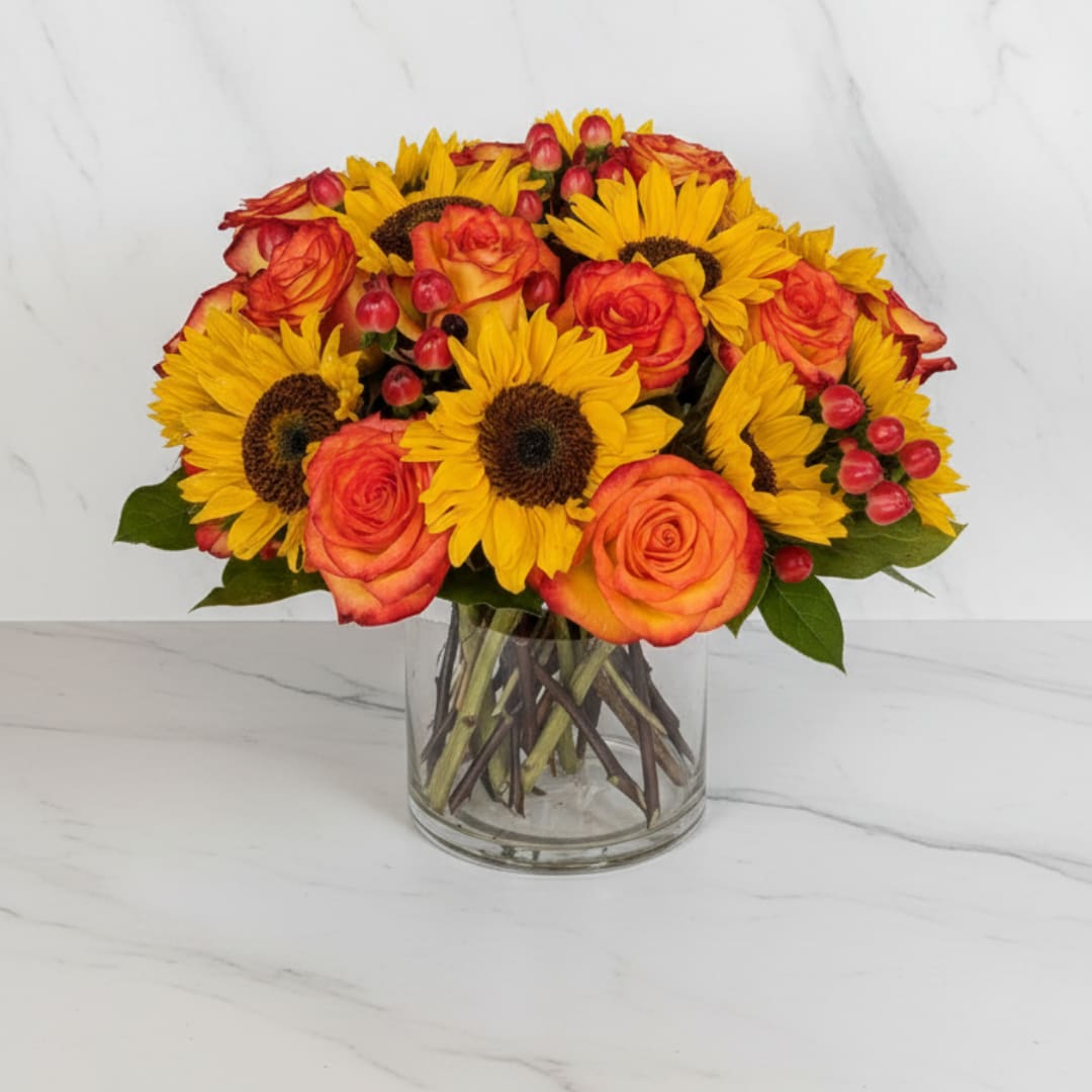 Harvest Embrace - A stunning seasonal favorite! Golden sunflowers and radiant orange roses are paired with berries and lush greens, arranged in a modern cylinder vase. Perfect for autumn gatherings, birthdays, or simply sending a heartfelt smile