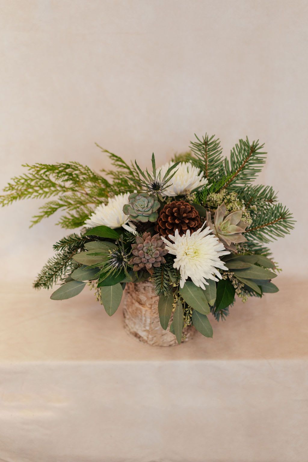Winters Magic - Seasonal greens, white flowers and blue thistle are arranged in a birch container. This is a great size for a centerpiece or kitchen table. Please read substitution policy.