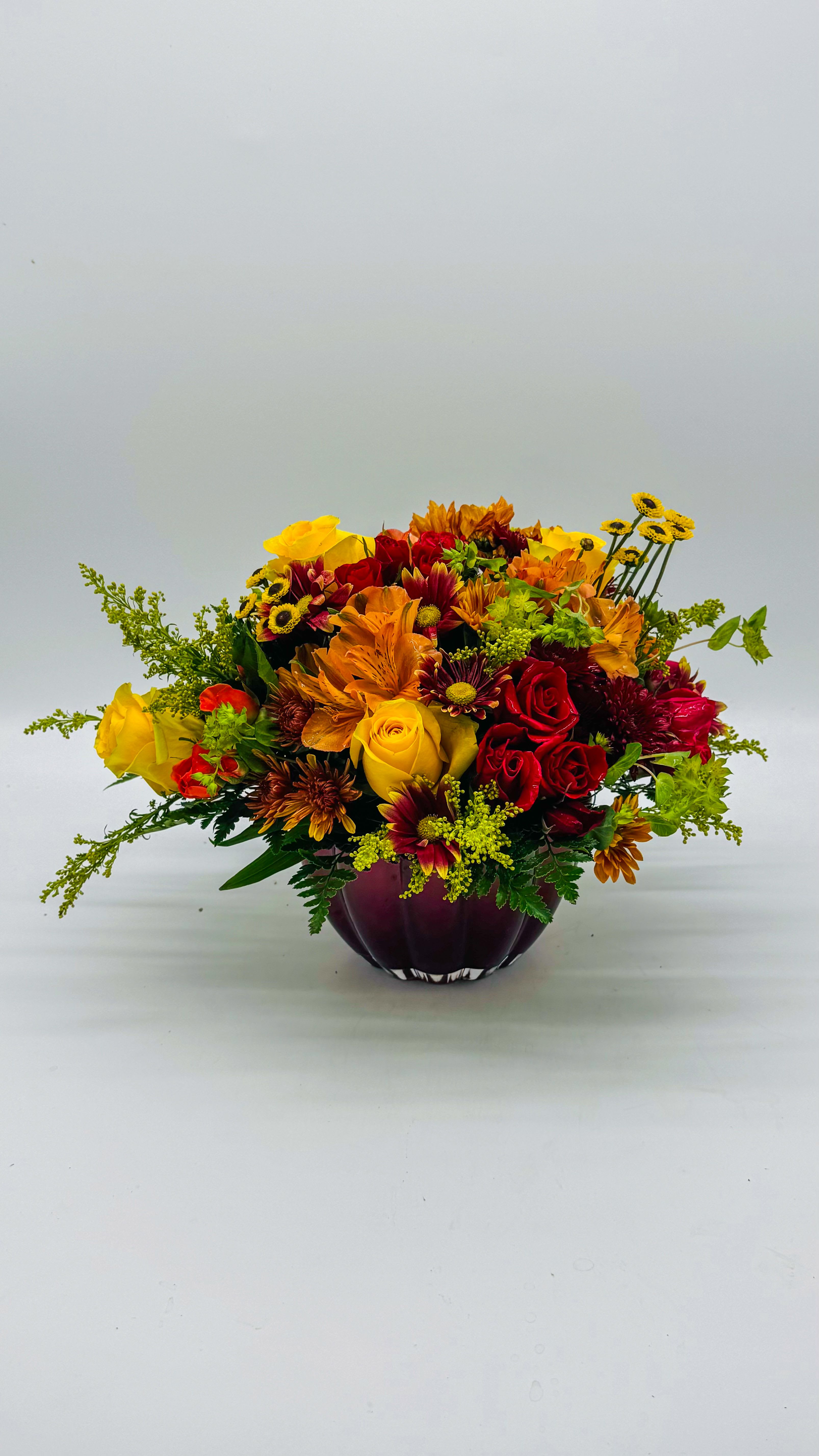 Crimson Harvest - A vibrant fall centerpiece crafted in a deep red bowl, this arrangement captures the warm glow of autumn. Rich crimson and burnt orange blooms mingle with soft amber centers, while sculpted bronze-green foliage adds depth and texture. Hints of dusky mauve and golden yellow echo the season’s sunsets, creating a cascade of color that feels both lush and grounded. The compact, balanced composition conveys cozy elegance, perfect for a tablescape or mantel to invite autumn’s charm into any room.