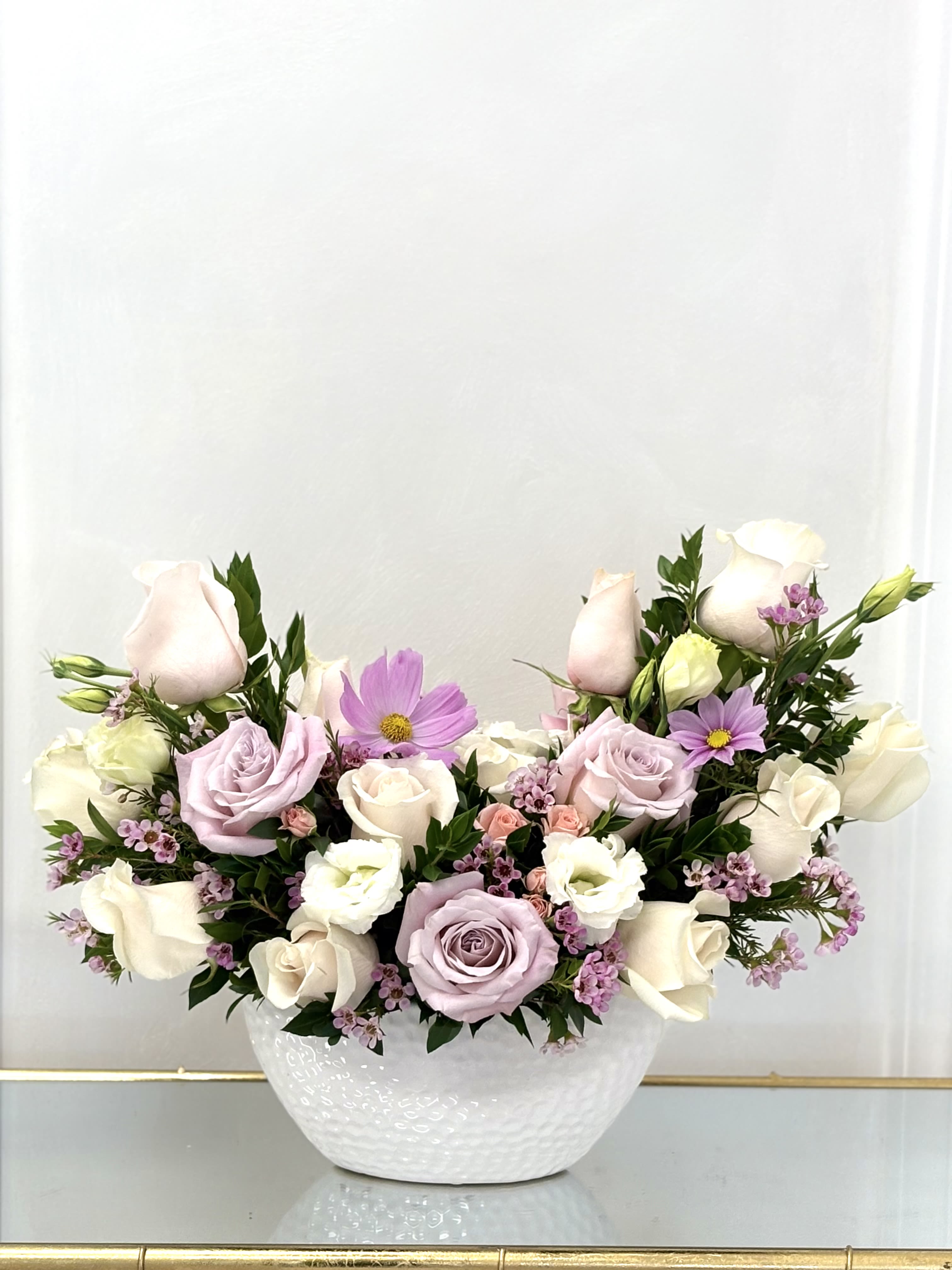 Blushing Meadow By VLM Flowers - Blushing Meadow is a soft and graceful arrangement that captures the spirit of a springtime garden. Featuring lavender roses, creamy white blooms, pale blush accents, and cheerful cosmos, this design is a gentle expression of love and serenity. Nestled in a textured white ceramic bowl, it radiates purity, elegance, and quiet joy — perfect for sympathy, a thoughtful gift, or a peaceful home accent.